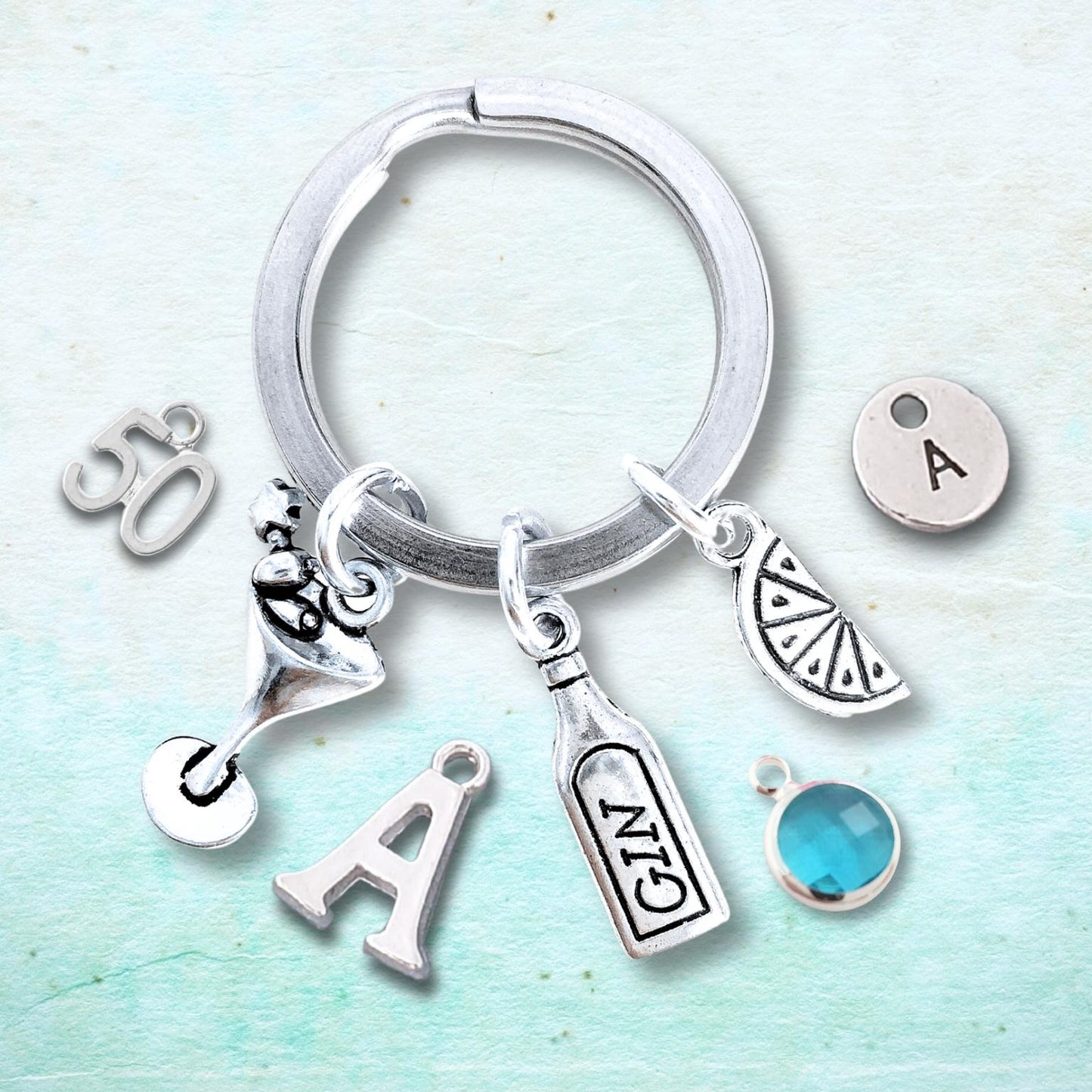 A silver-toned key ring charm set featuring a martini glass, gin bottle, citrus slice, letter 'A', number '50', and an initial 'A' charm with a turquoise blue gemstone, displayed on a textured light blue background.