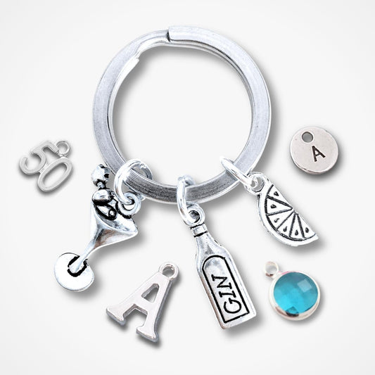 A silver keychain ring with multiple charms, including a martini glass, a gin bottle, a citrus slice, a letter 'A', a '50' charm, a letter 'A' initial, and a round turquoise blue gemstone charm, displayed on a light gray background.