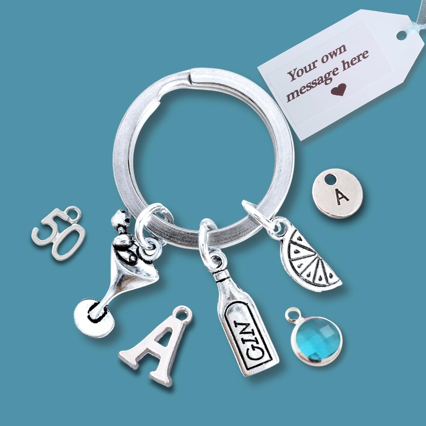 A silver keychain with multiple charms, including an initial 'A', a martini glass, a bottle, a slice of citrus, a round letter 'A' charm, and a blue faceted gem, displayed on a solid teal background with a white customization tag.