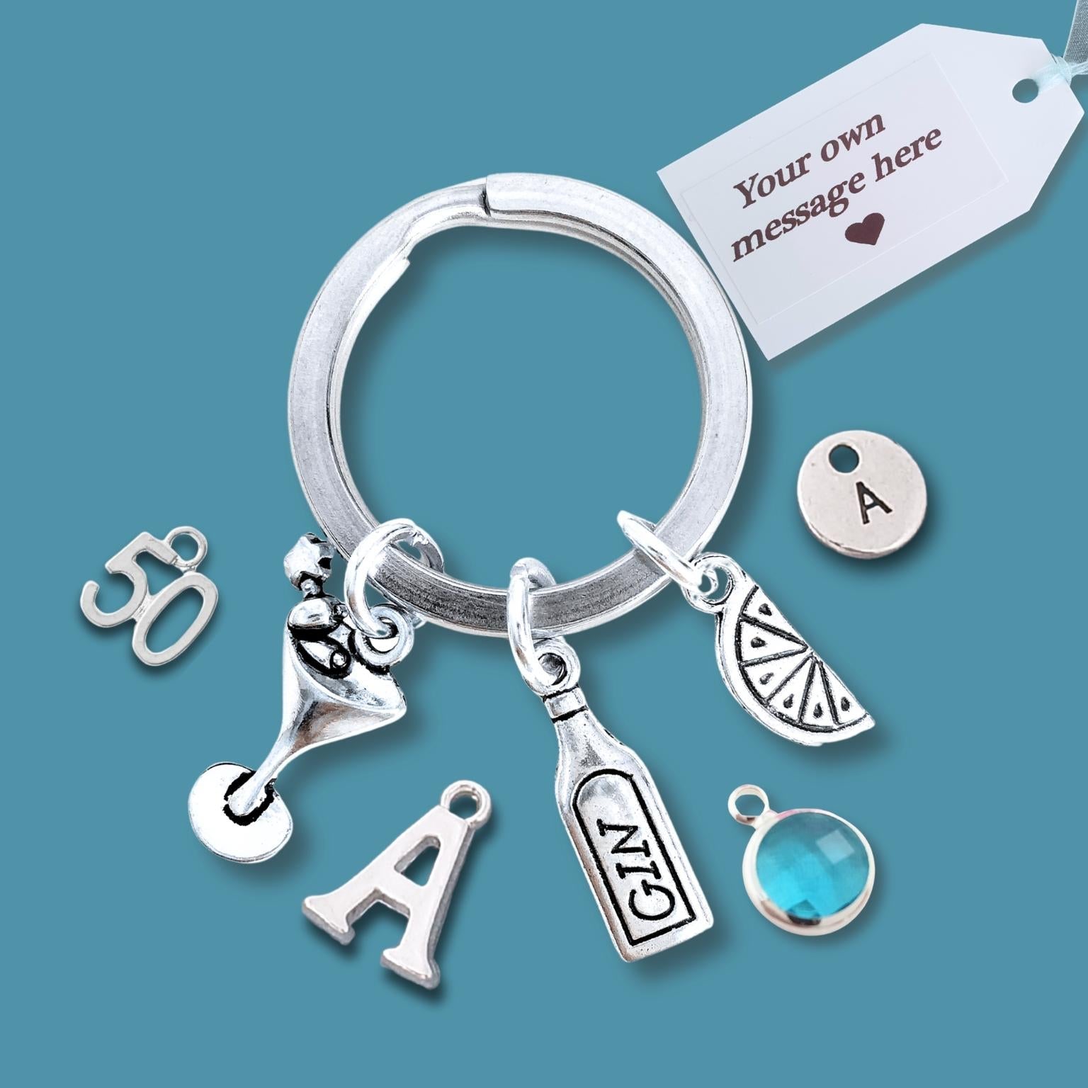A silver keychain with multiple charms, including an initial 'A', a martini glass, a bottle, a slice of citrus, a round letter 'A' charm, and a blue faceted gem, displayed on a solid teal background with a white customization tag.