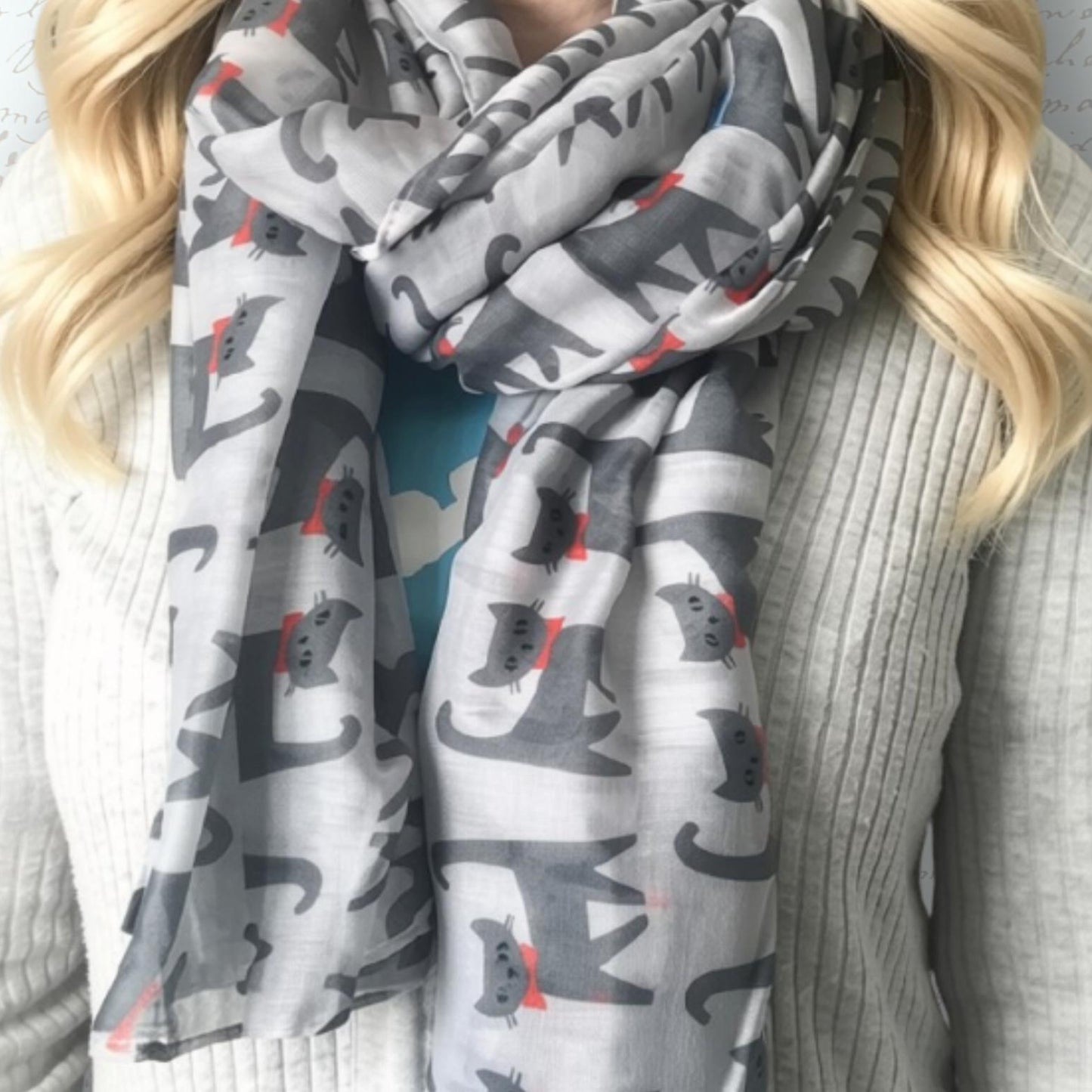 A lightweight grey scarf with a repeating pattern of dark grey cats and small red bows, worn by a blonde woman in a ribbed light grey sweater.