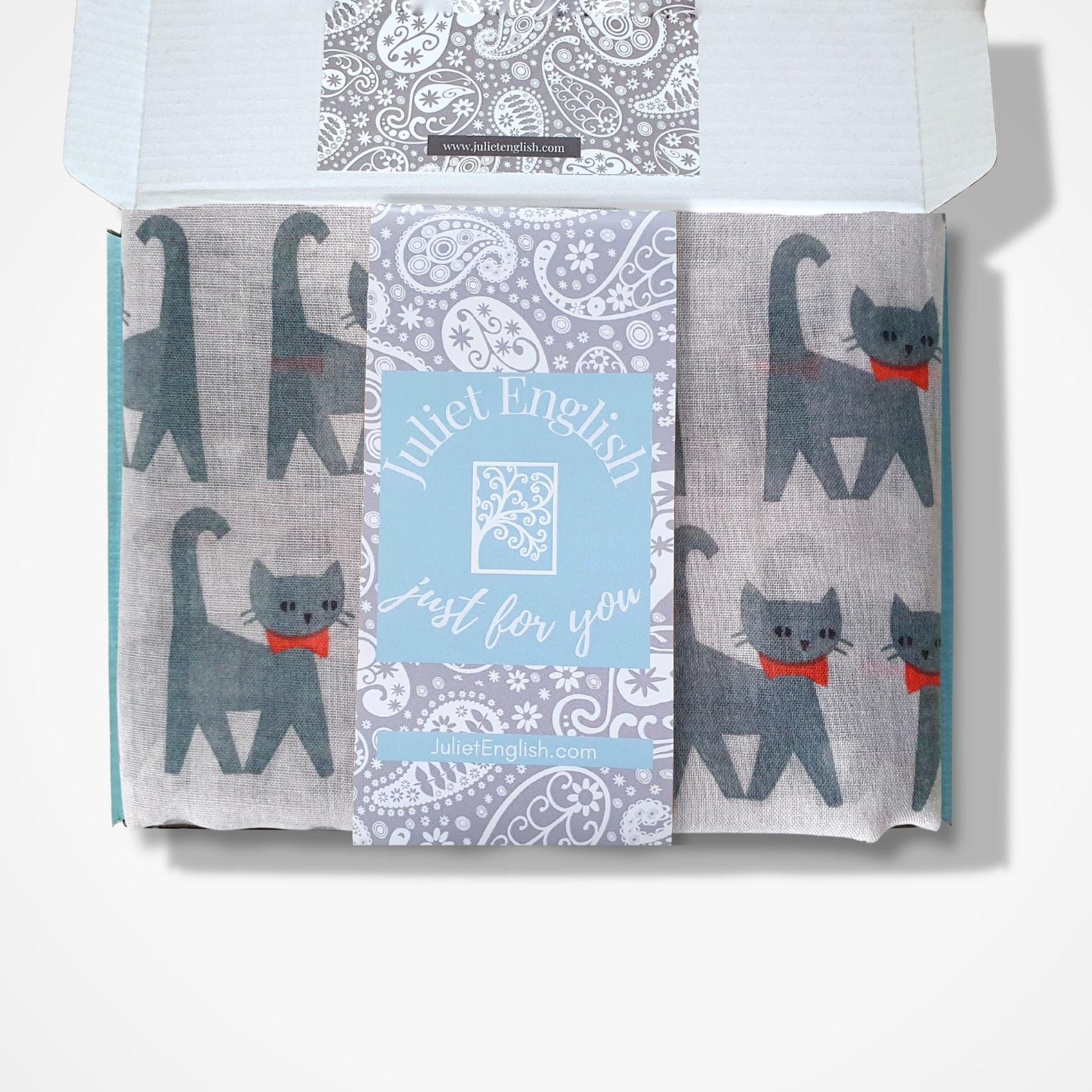 A boxed scarf from Juliet English, featuring grey cat illustrations with red bows and a central paisley pattern, displayed in an open box against a white background.