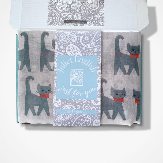 A boxed scarf from Juliet English, featuring grey cat illustrations with red bows and a central paisley pattern, displayed in an open box against a white background.