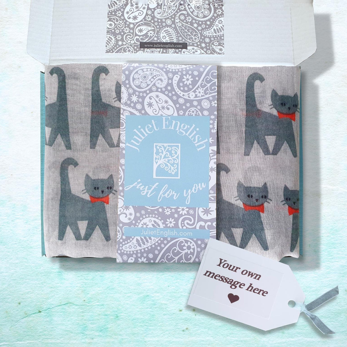 A gift box containing a folded scarf with grey cat illustrations and a central blue label reading 'Juliet English Just for you,' plus a small white tag that says 'Your own message here' on a pale aqua textured background.