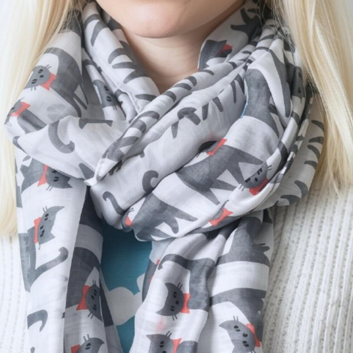 A close-up of a person wearing a lightweight grey scarf with a playful print of dark grey cats and small red bows, paired with a grey ribbed knit sweater.