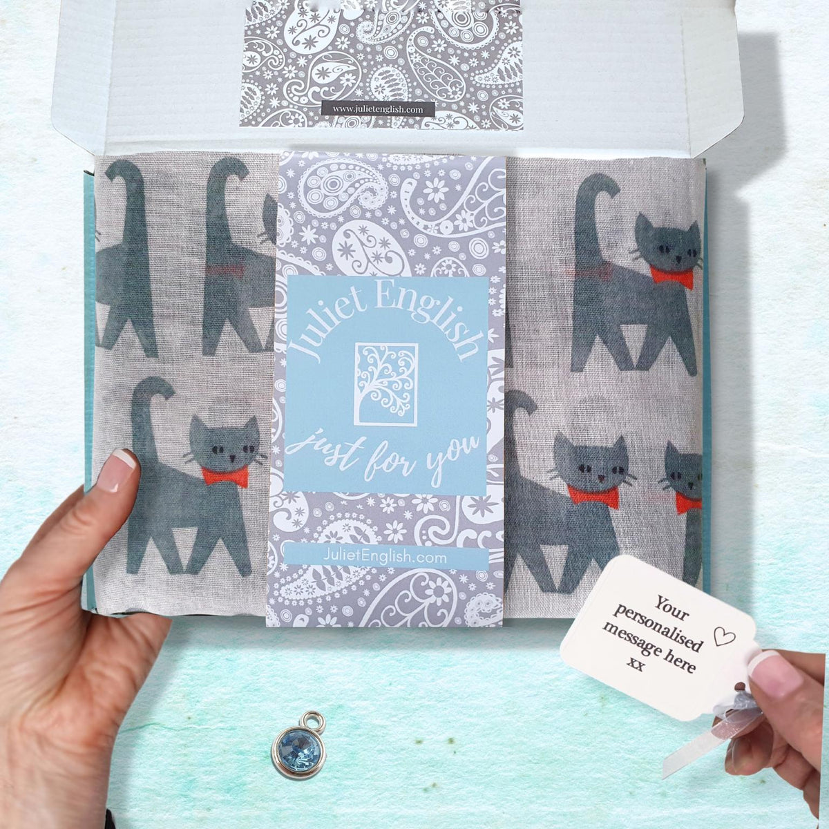 A hand holds an open white gift box containing a scarf with grey cats and red bowties, a light blue central card reading 'Juliet English Just for you,' and a small white tag. A small round pendant lies below the box on a pale blue textured surface.