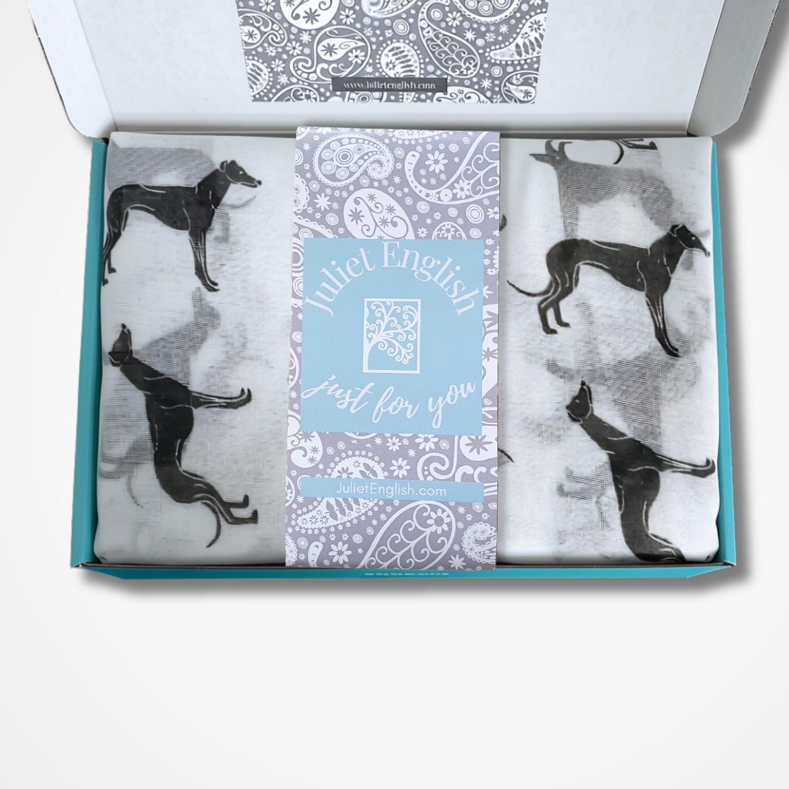 An open gift box containing a scarf with black greyhound silhouettes on a white background, plus a central light blue card that reads 'Juliet English Just for you'.