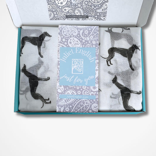 An open gift box containing a scarf with black greyhound silhouettes on a white background, plus a central light blue card that reads 'Juliet English Just for you'.