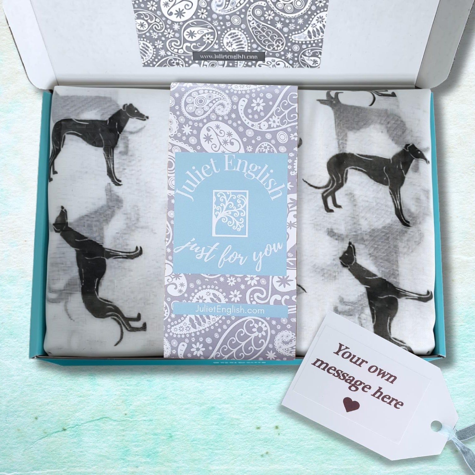 An open gift box containing a scarf with black greyhound silhouettes on a white background, plus a central light blue card that reads 'Juliet English Just for you' and a white gift tag that reads 'Your own message here'.