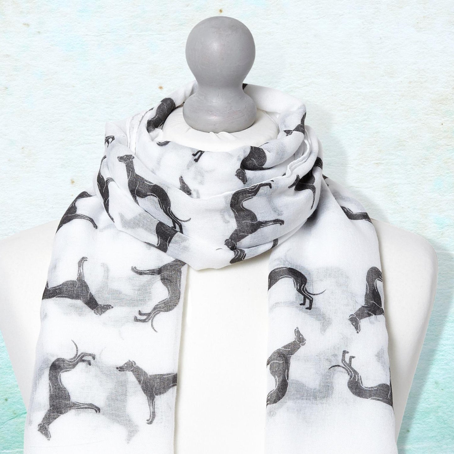 A scarf with black greyhound silhouettes on a white background, draped on a white mannequin torso against a textured light blue background.