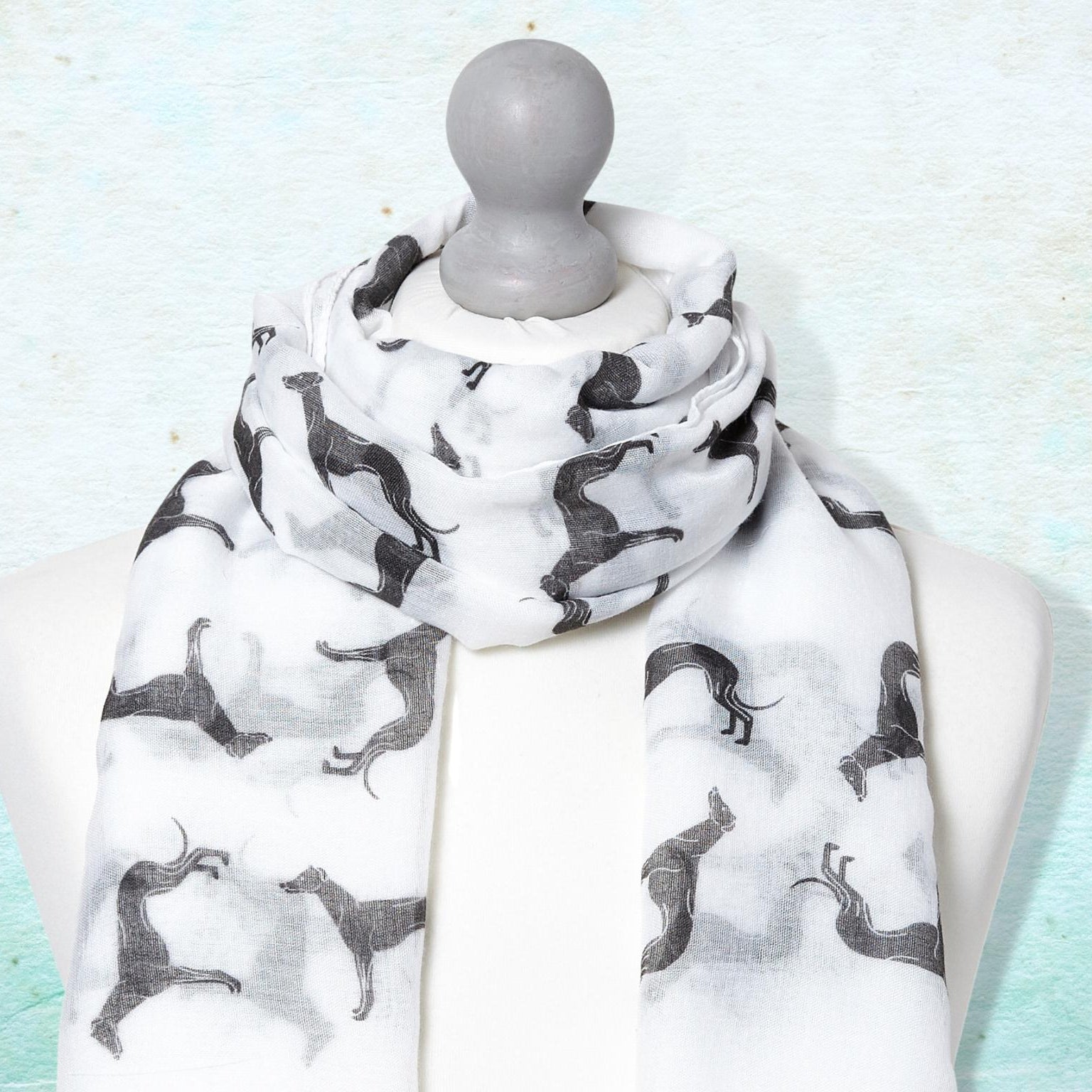 A scarf with black greyhound silhouettes on a white background, draped on a white mannequin torso against a textured light blue background.