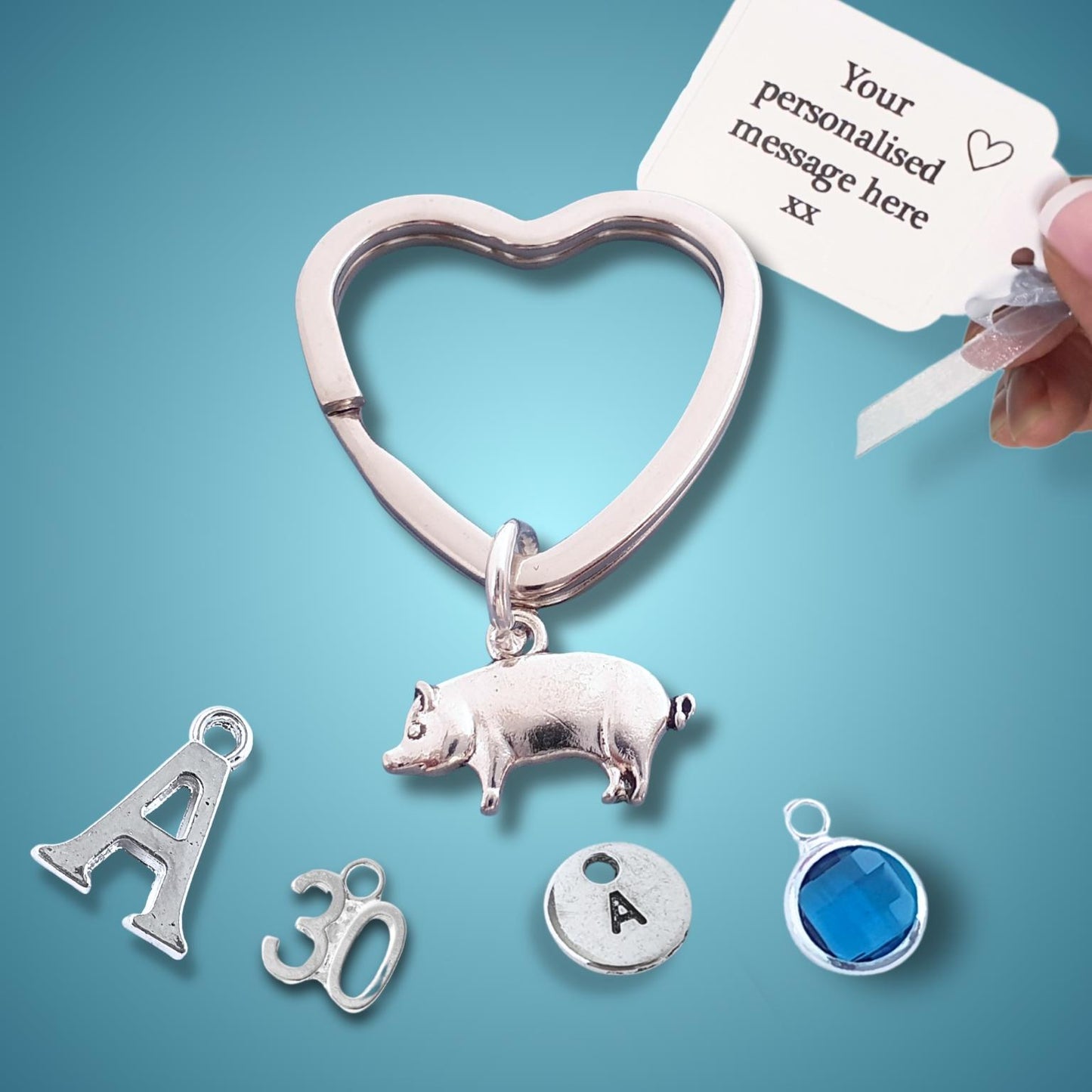 Heart-shaped pig keychain with charms and a hand holding a card for personalization on a blue background