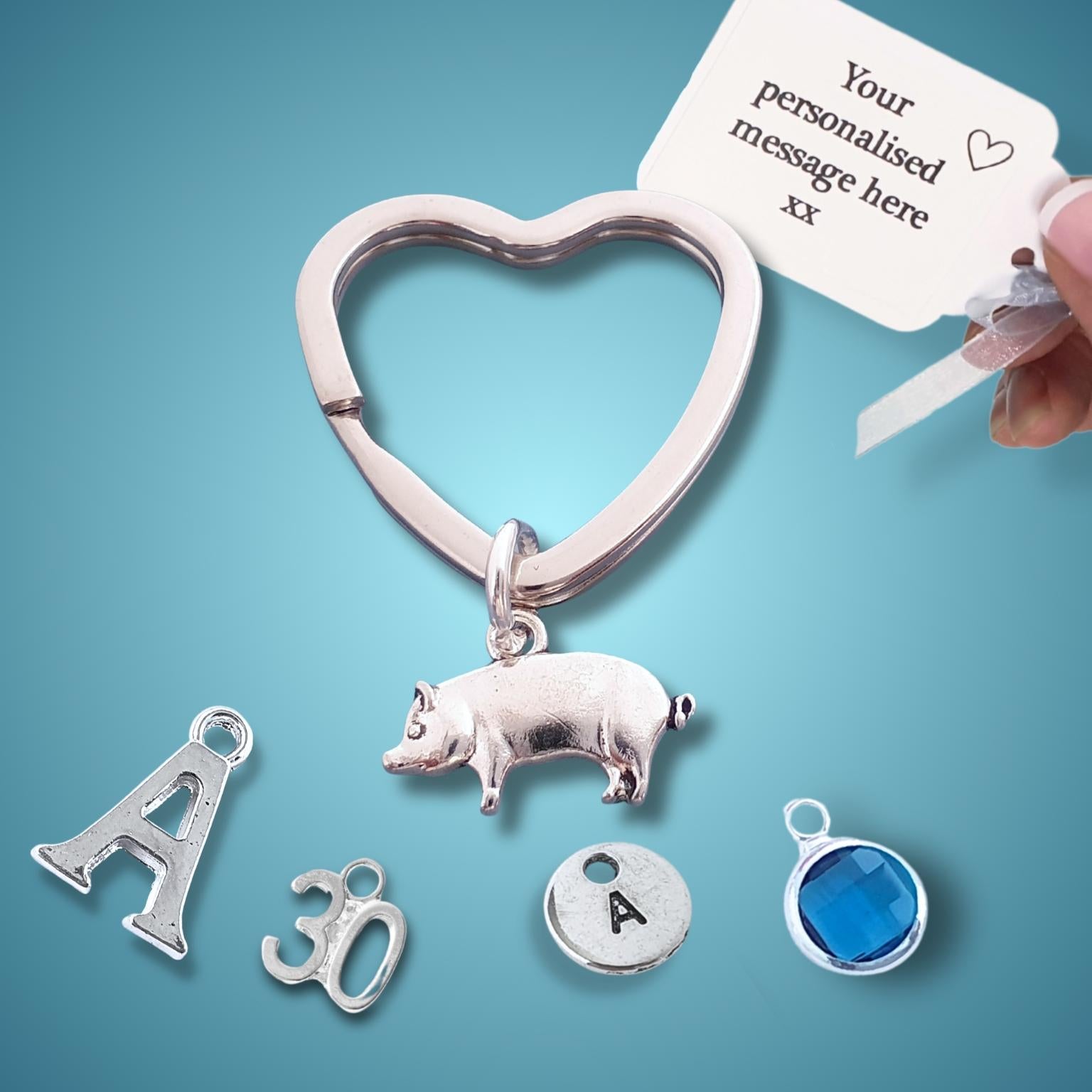 Heart-shaped pig keychain with charms and a hand holding a card for personalization on a blue background