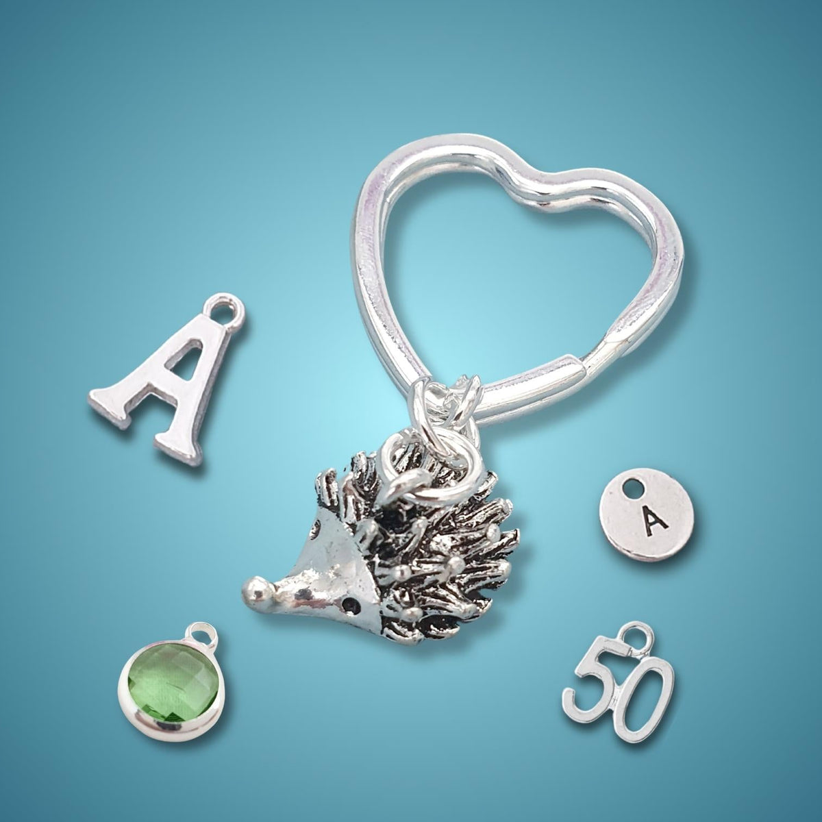 A silver keychain with a heart-shaped ring, a hedgehog charm, and several customizable charms including an 'A' letter, a round 'A' initial, a green gemstone bezel, and a '50' charm, displayed on a blue gradient background.
