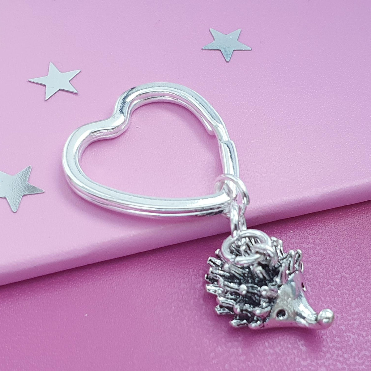 A silver heart-shaped key ring with a small hedgehog charm, displayed on a pink background with star-shaped confetti.
