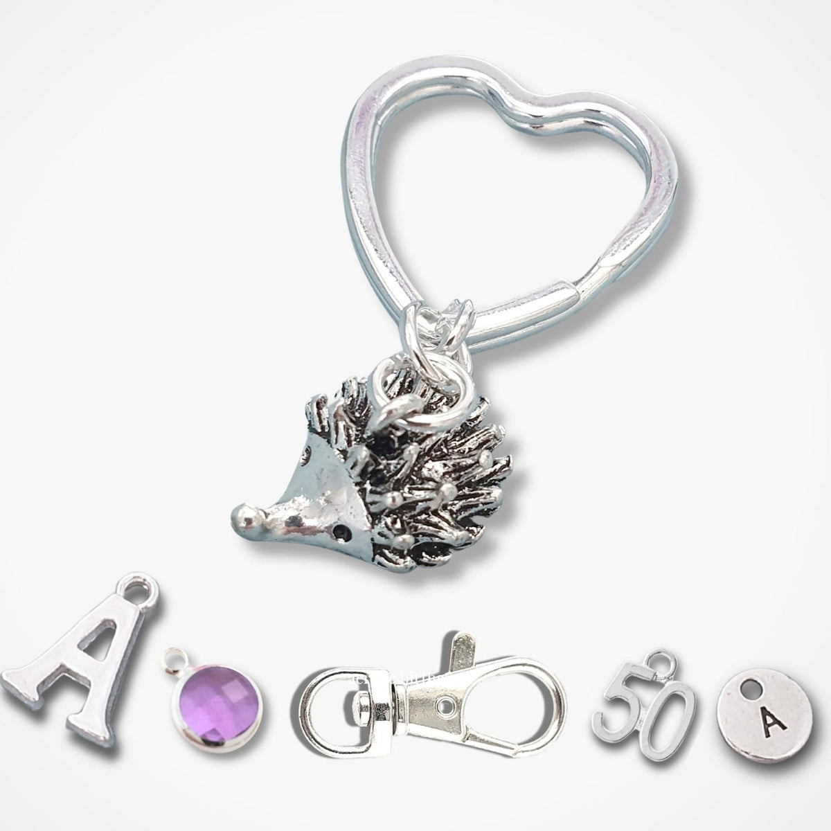 Silver metal keyring featuring a hedgehog charm, a heart-shaped ring, a lobster clasp, and small letter and gemstone charms on a light gray background.