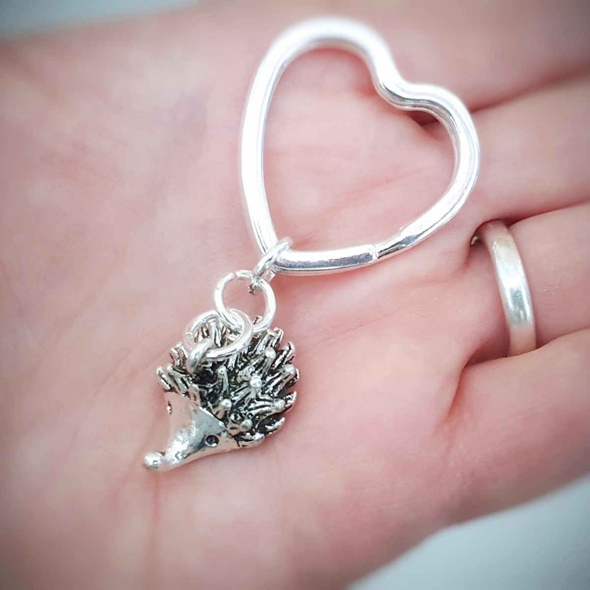A sterling silver heart-shaped ring with a dangling hedgehog charm, held in a person's hand.
