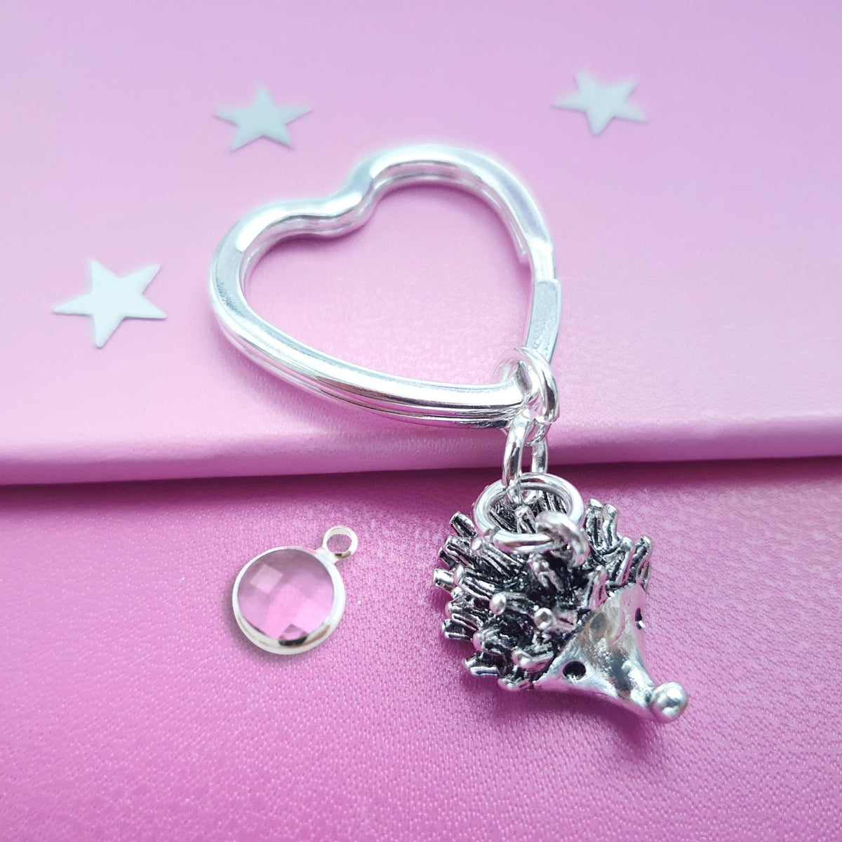 A silver heart-shaped keyring with a dangling hedgehog charm, displayed on a pink background with a small separate silver ring and clear gemstone.