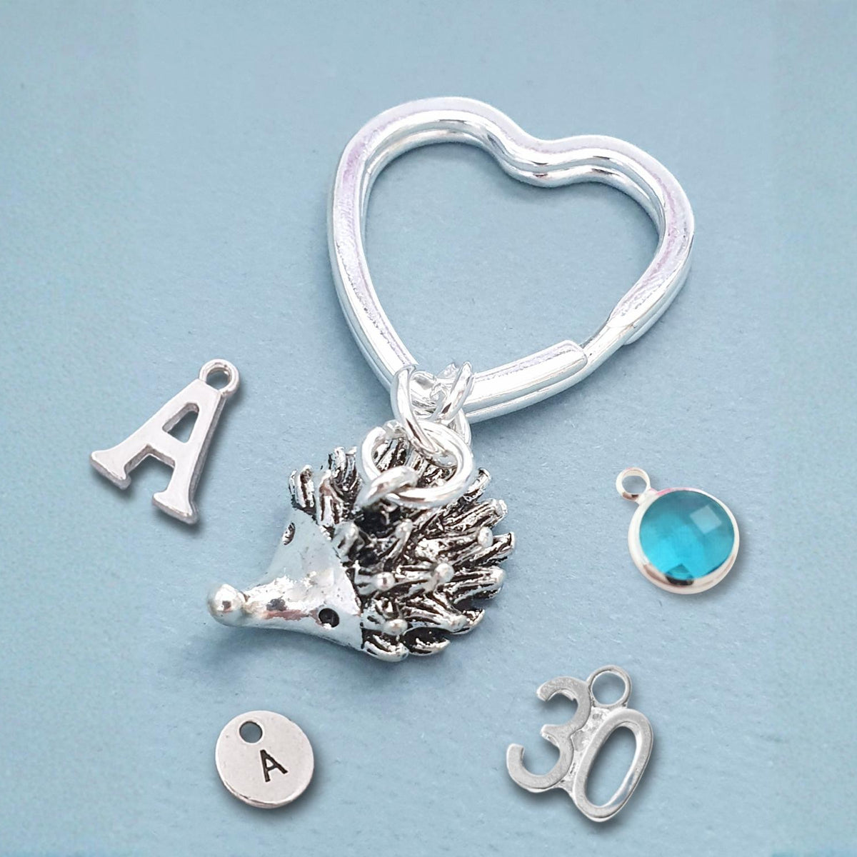 A silver heart-shaped key ring charm set displayed on a light blue background, featuring a textured hedgehog charm, a letter 'A' initial charm, a '30' charm, a round silver disc with the letter 'A', and a small round turquoise blue cabochon charm.