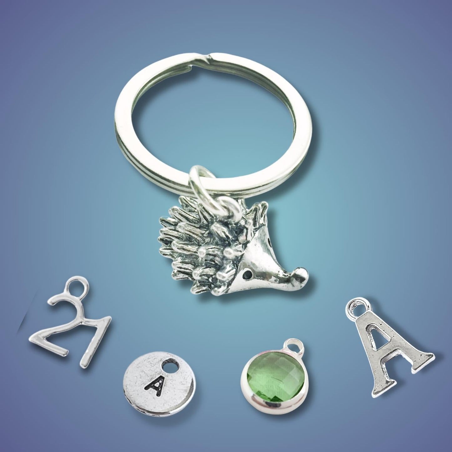 Silver hedgehog keychain with various charms including a number 21, letter A, and green gemstone on a blue gradient background.