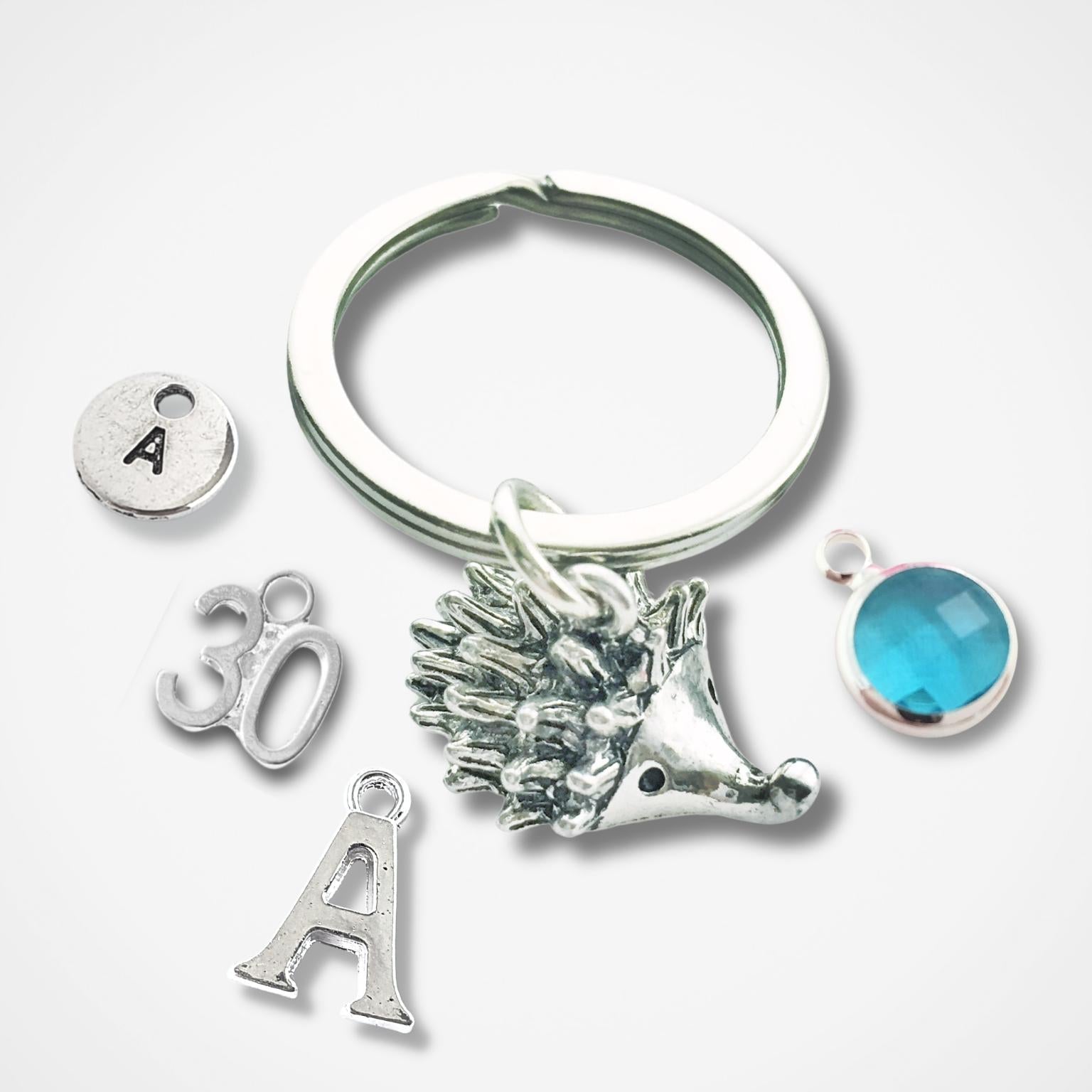 Silver hedgehog keychain with various charms including a hand, letter 'A', and blue stone on a white background