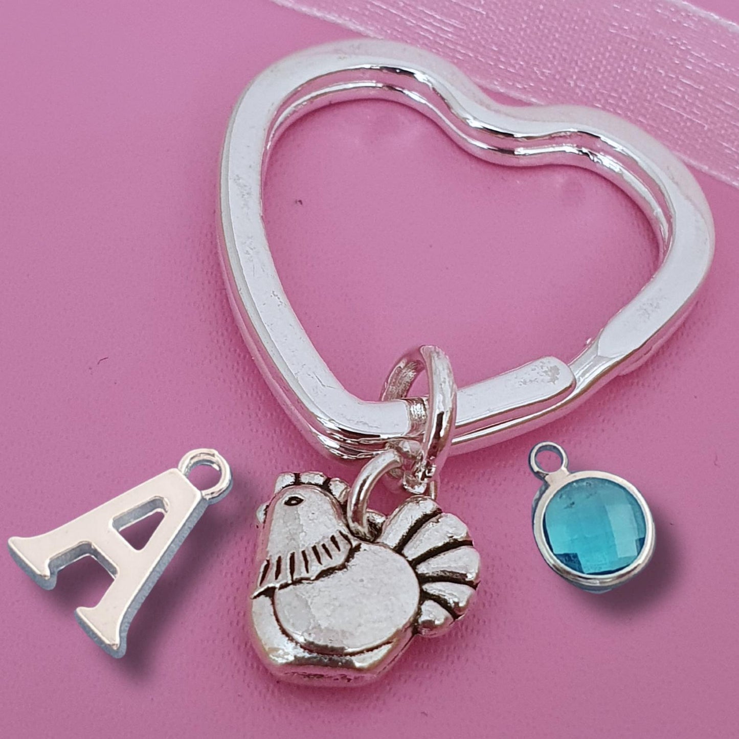 A silver-toned heart-shaped key ring charm with three dangling charms: a letter 'A', a hen, and a round blue faceted gemstone, displayed on a pink background.