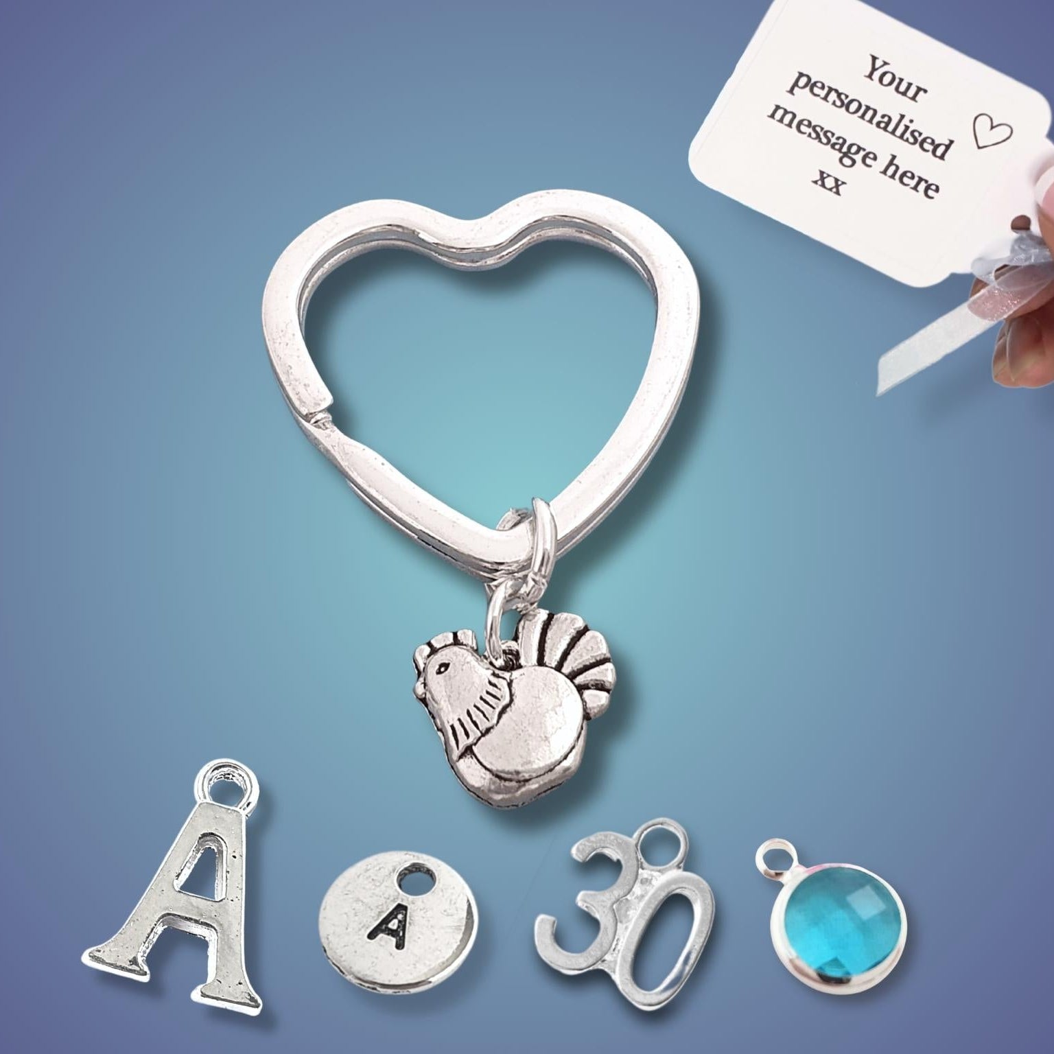 A silver heart-shaped key ring with a hen pendant and individual letter/number/initial charms, shown on a blue gradient background with a hand holding a customization tag.