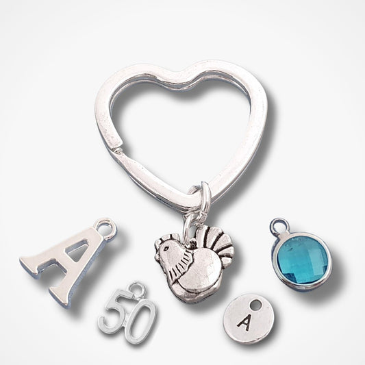 A silver heart-shaped hen key ring charm with dangling personalized charms, including a letter 'A', the number '50' and a round bezel-set blue stone charm, shown on a light gray background.