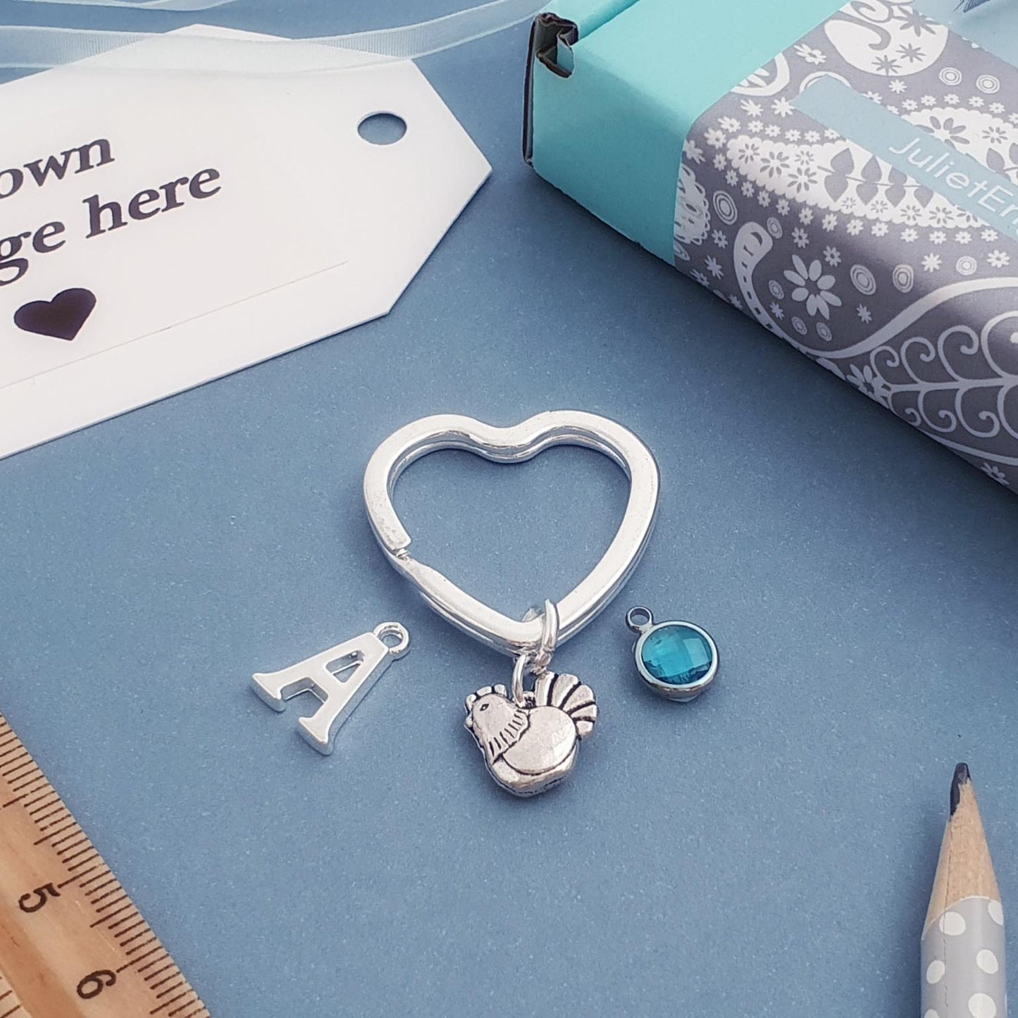 A silver heart-shaped charm keychain with a letter 'A' pendant, a hen charm, and a small turquoise stone charm, displayed on a blue background with a gift tag, ruler, pencil, and gift box.