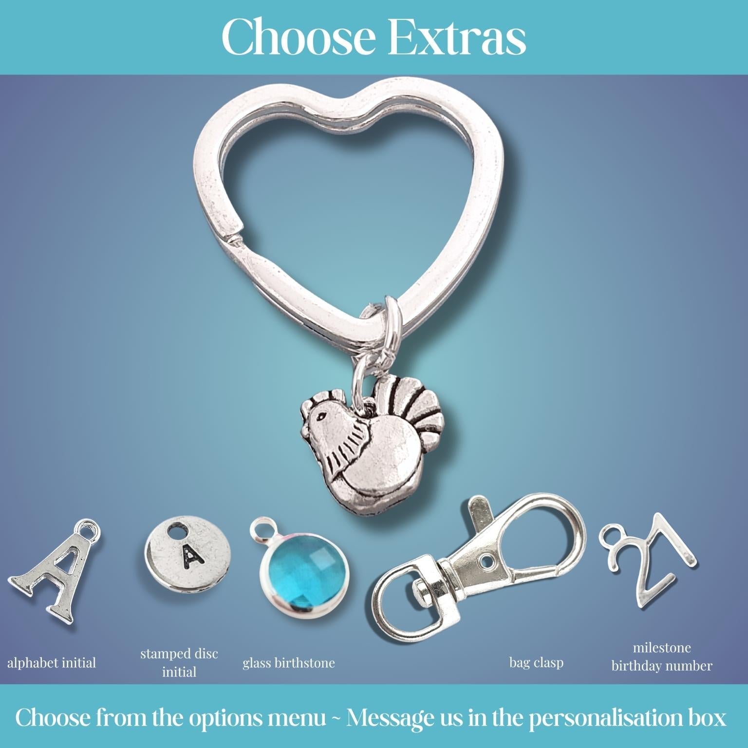 A promotional graphic showing a silver heart-shaped clasp with a small hen charm, plus several customizable extra options labeled alphabet initial, stamped date initial, glass birthstone, bag charm, and milestone birthday number on a blue gradient background with the heading 'Choose Extras'.