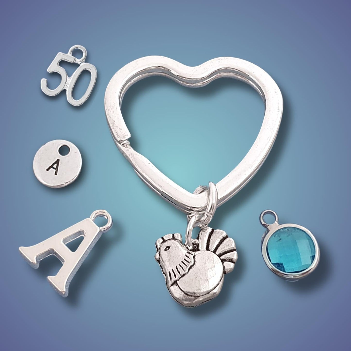 A heart-shaped keyring with a hen charm, an 'A' initial charm, a '50' charm, a small round charm with the letter 'A' and a blue faceted gemstone bezel.