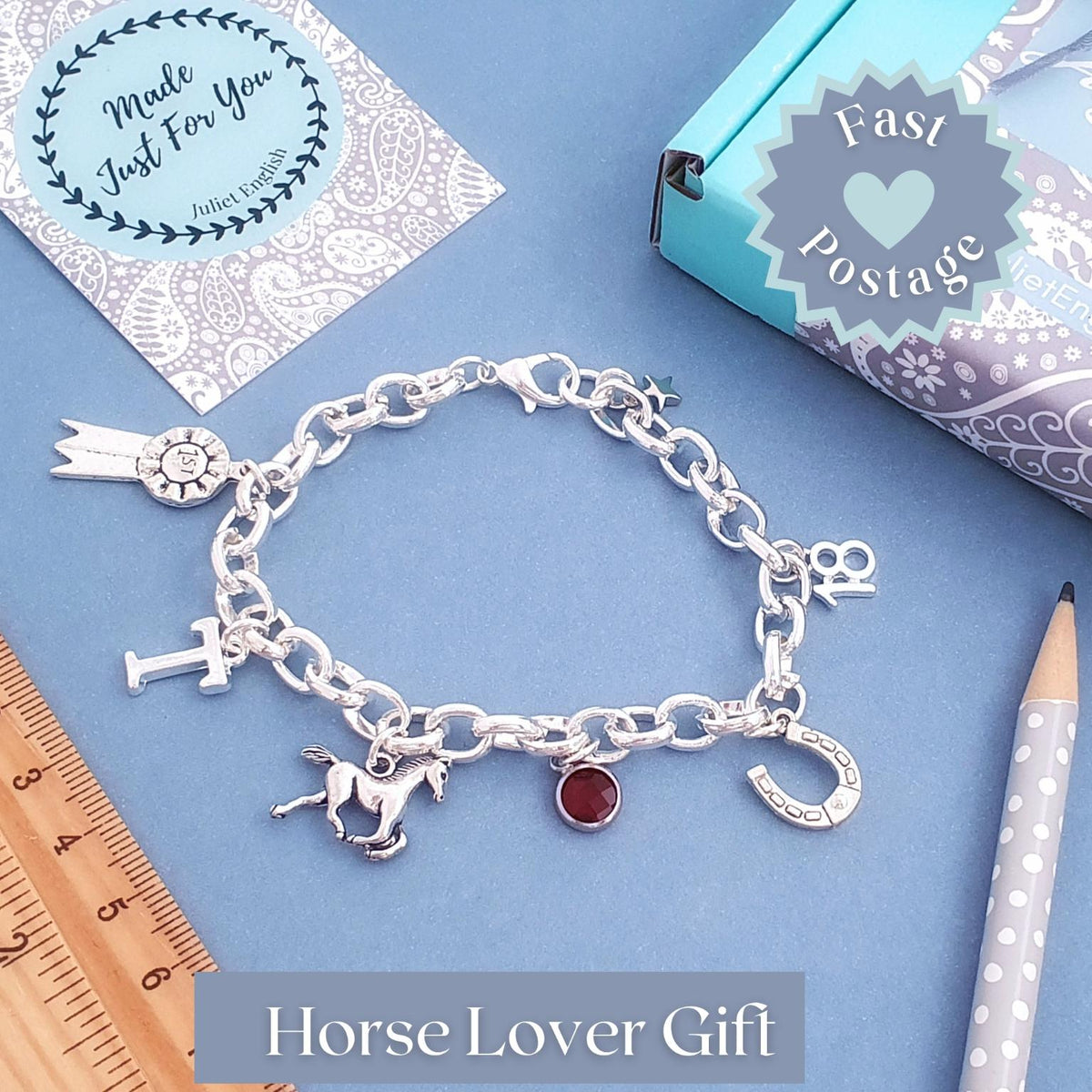 A silver charm bracelet with horse-themed charms, including a horse silhouette, horseshoe, and rosette, displayed on a blue background with a wooden ruler, pencil, and gift packaging.