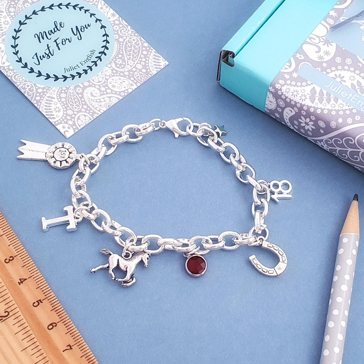 A silver charm bracelet with equestrian-themed charms including a horse, horseshoe, and rosette, displayed on a blue surface with a wooden ruler, pencil, and gift box.