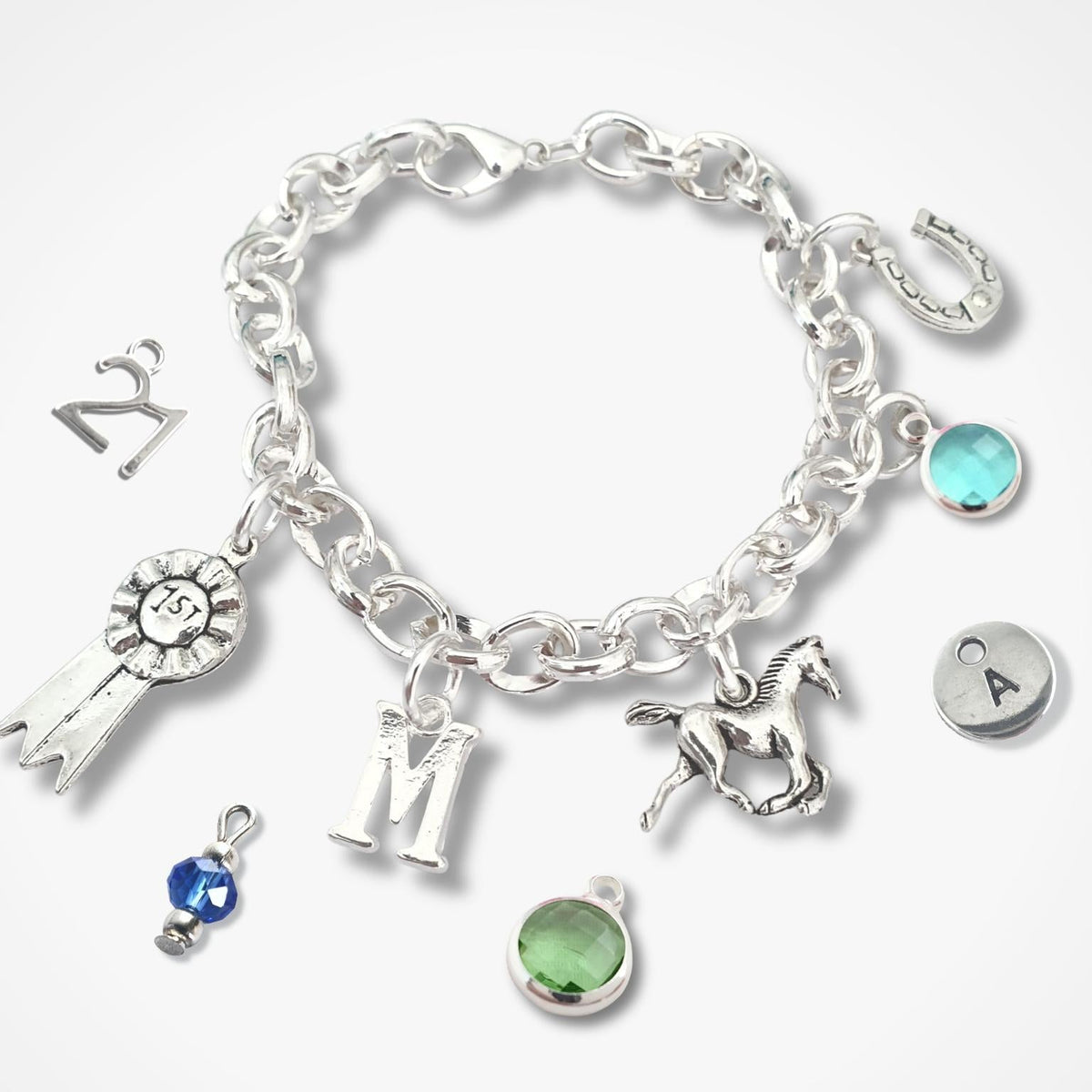 A silver charm bracelet with multiple dangling charms, including a horse, rosette ribbon, letter 'M', horseshoe, and round gemstone charms, displayed on a light gray background.