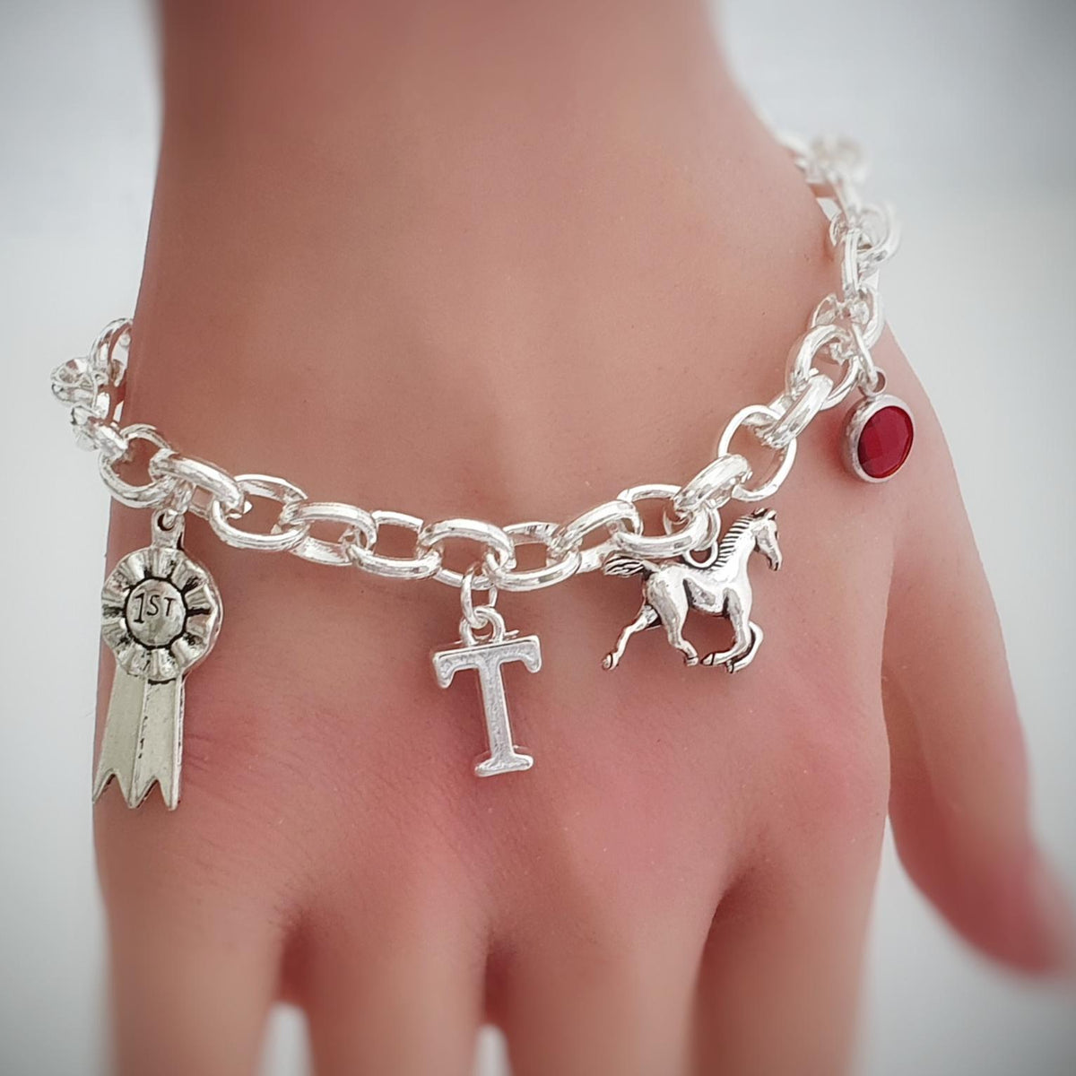 A close-up of a hand wearing a silver charm bracelet with horse and rosette ribbon charms, set against a soft, blurred gray background.