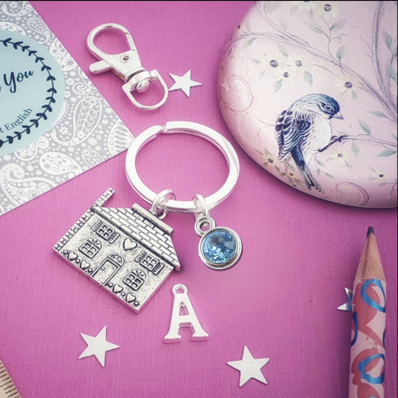 Keychain with house charm, letter 'A' charm, and blue gemstone on a pink surface with decorative elements.