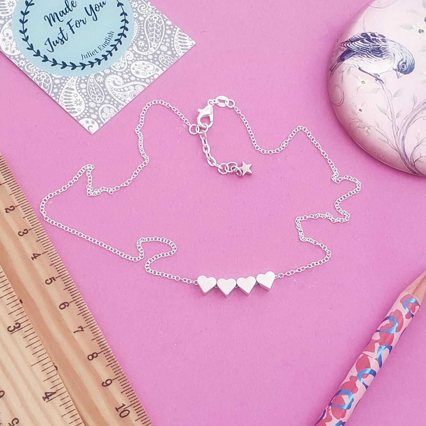 Silver necklace with heart pendants on a pink background with a ruler and pencil.