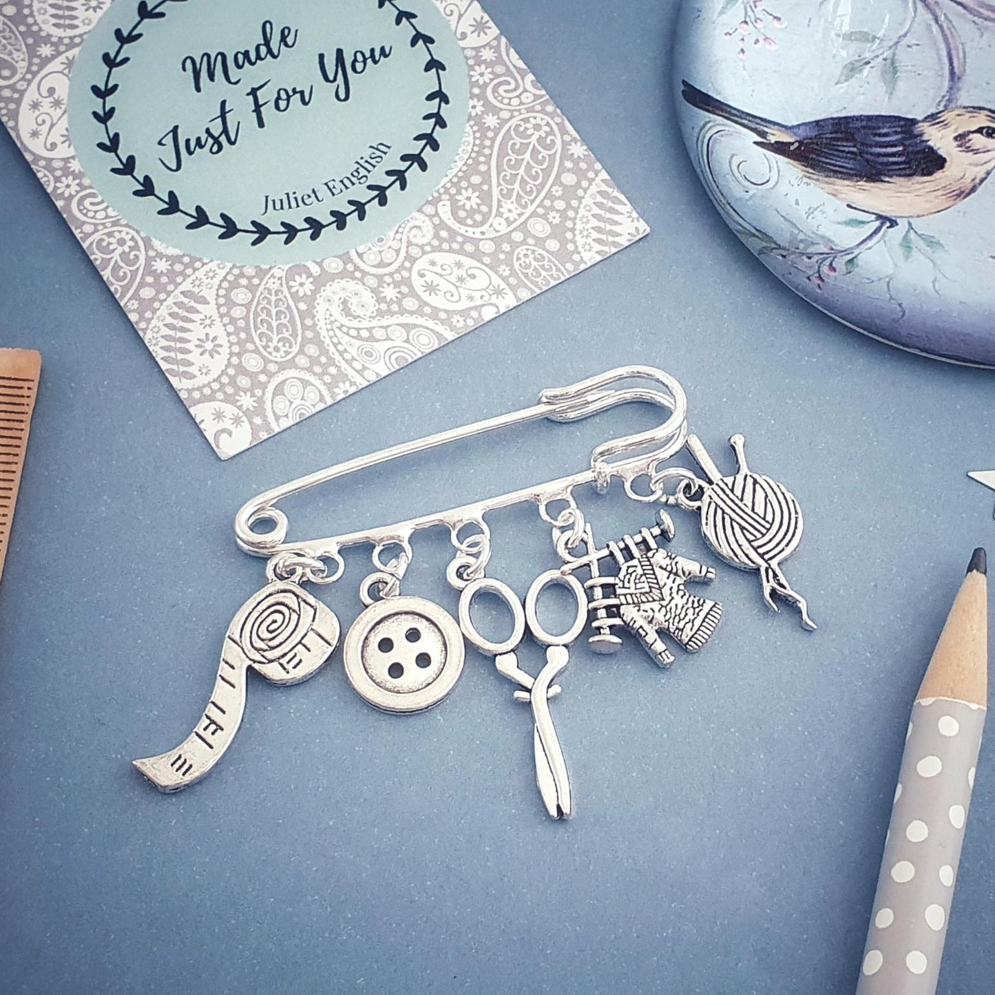Silver brooch with various knitting themed charms on a blue surface with a card and pencil.