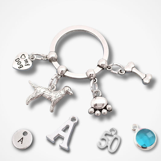 A silver-tone charm key ring with multiple dangling charms, including a dog silhouette, paw print, bone, heart with 'Ch My Dog' text, and letter/number charms, shown on a light gray background with additional loose charms below.