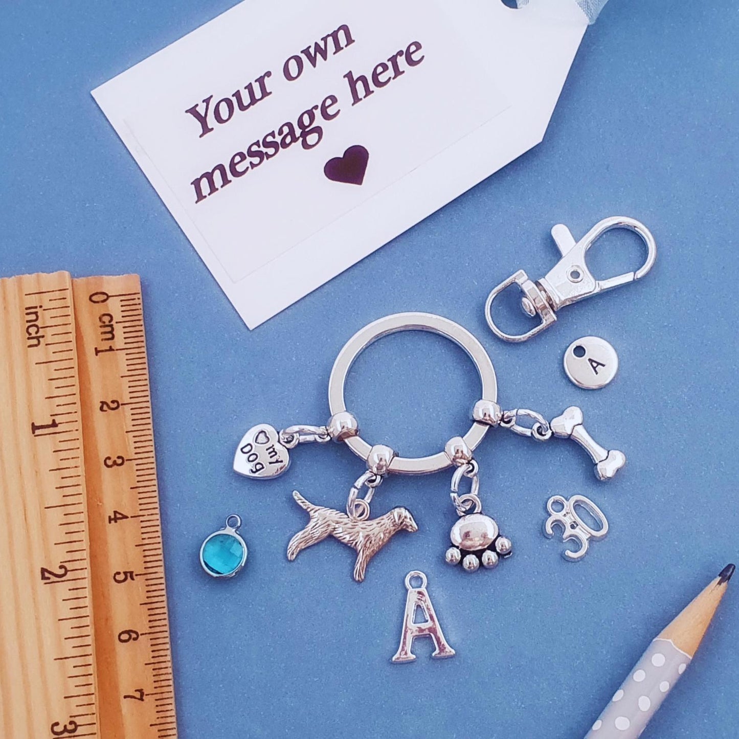 A silver-toned keyring charm set displayed on a blue background, including a dog silhouette charm, paw print, bone, heart, and letter 'A' charms, alongside a clasp, tag, ruler, and pencil.