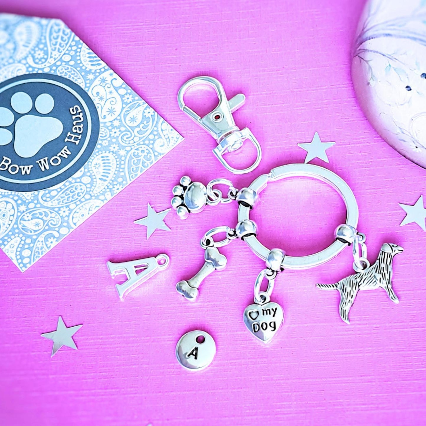 A silver pet-themed charm key ring set displayed on a bright pink background, featuring a dog silhouette charm, a heart charm engraved with 'my dog', a paw print charm, and a small round charm with the letter 'A'. A Bow Wow Haus branded card is visible in the top left corner.