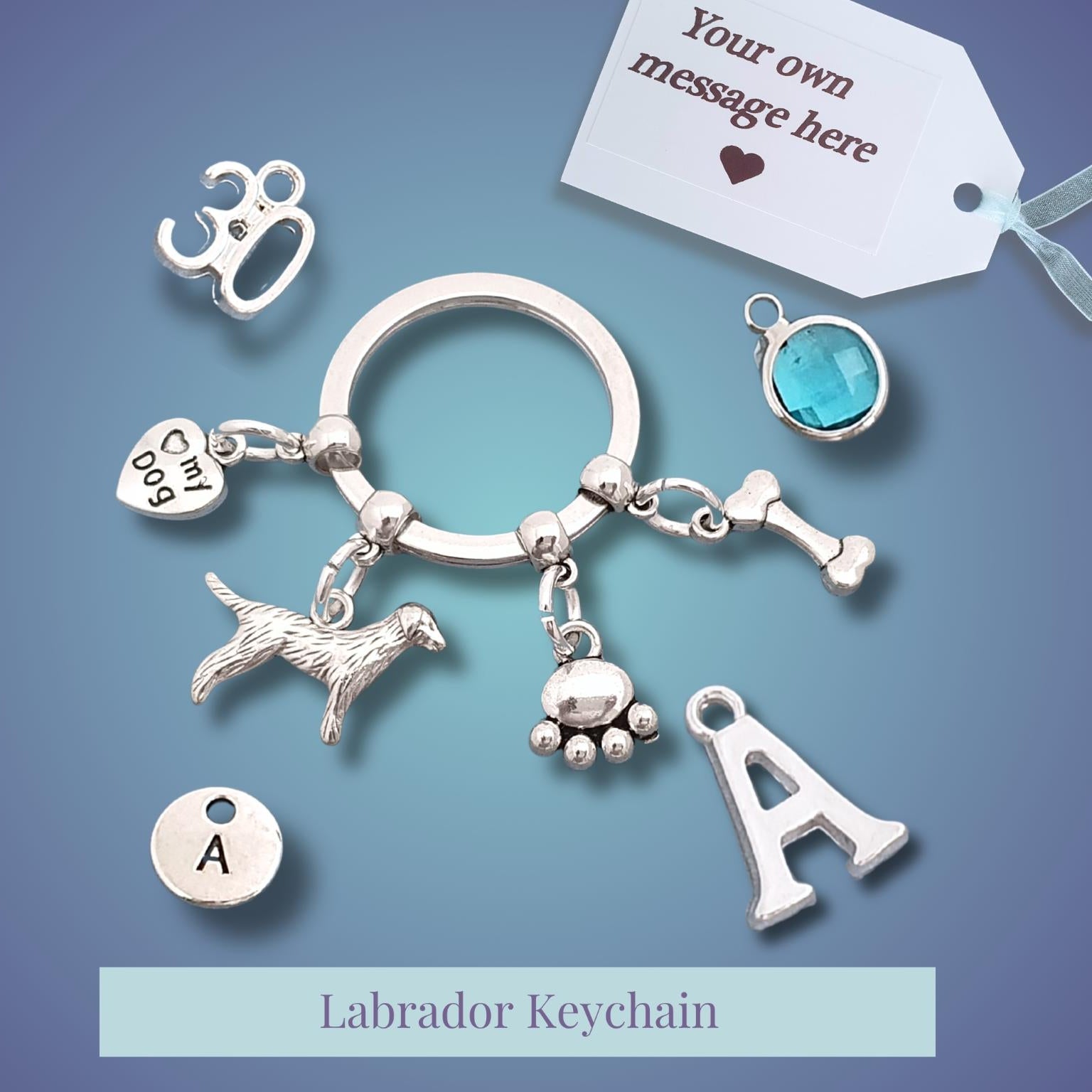 Silver-tone Labrador keychain charm set with multiple dangling charms including a dog silhouette, paw print, bone, letter 'A', and heart-shaped tags, displayed on a blue gradient background with a customization tag and text label.
