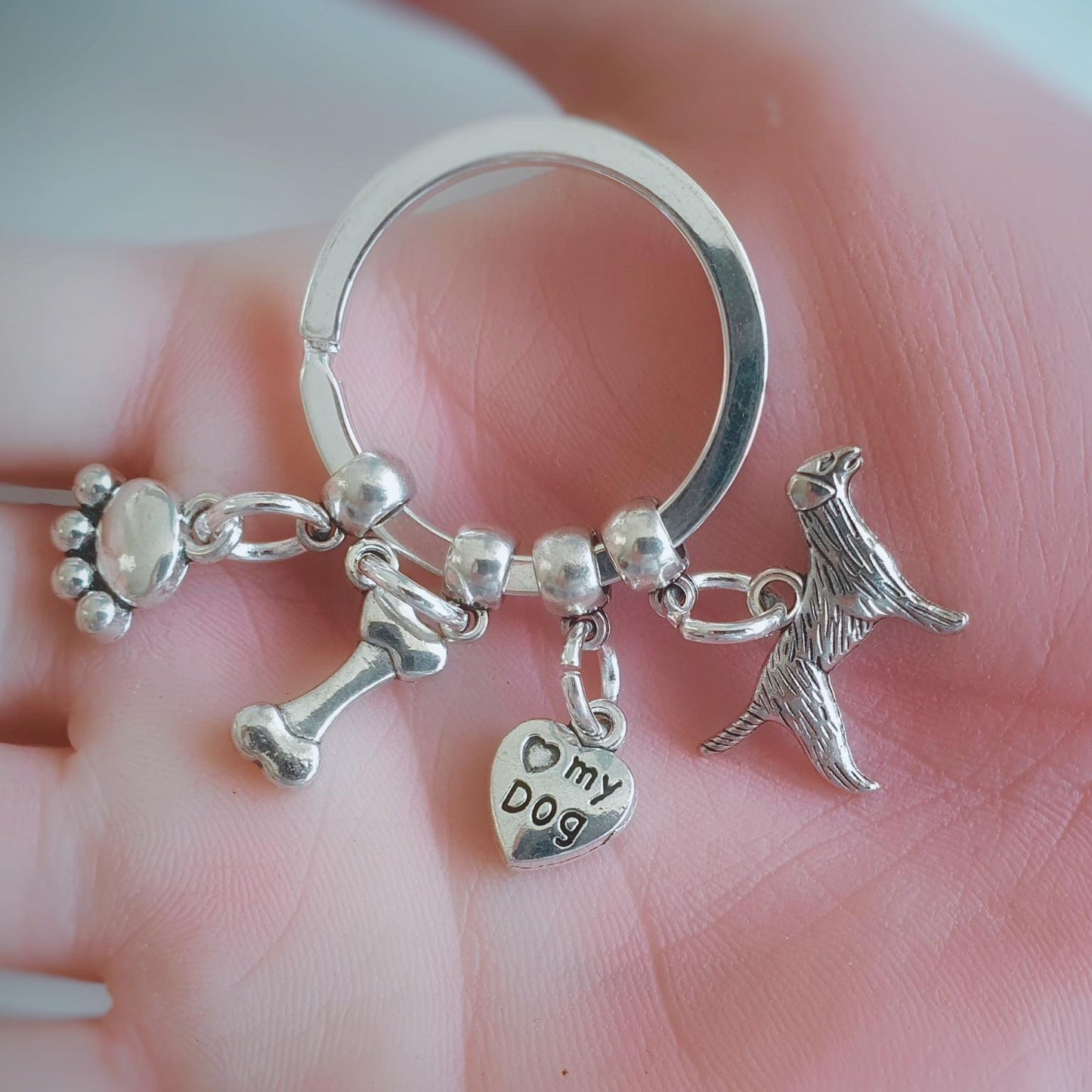 A silver-tone key ring charm set held in a person's hand. The set includes a paw print charm, a bone charm, a heart charm stamped with 'my dog', and a dog silhouette charm.