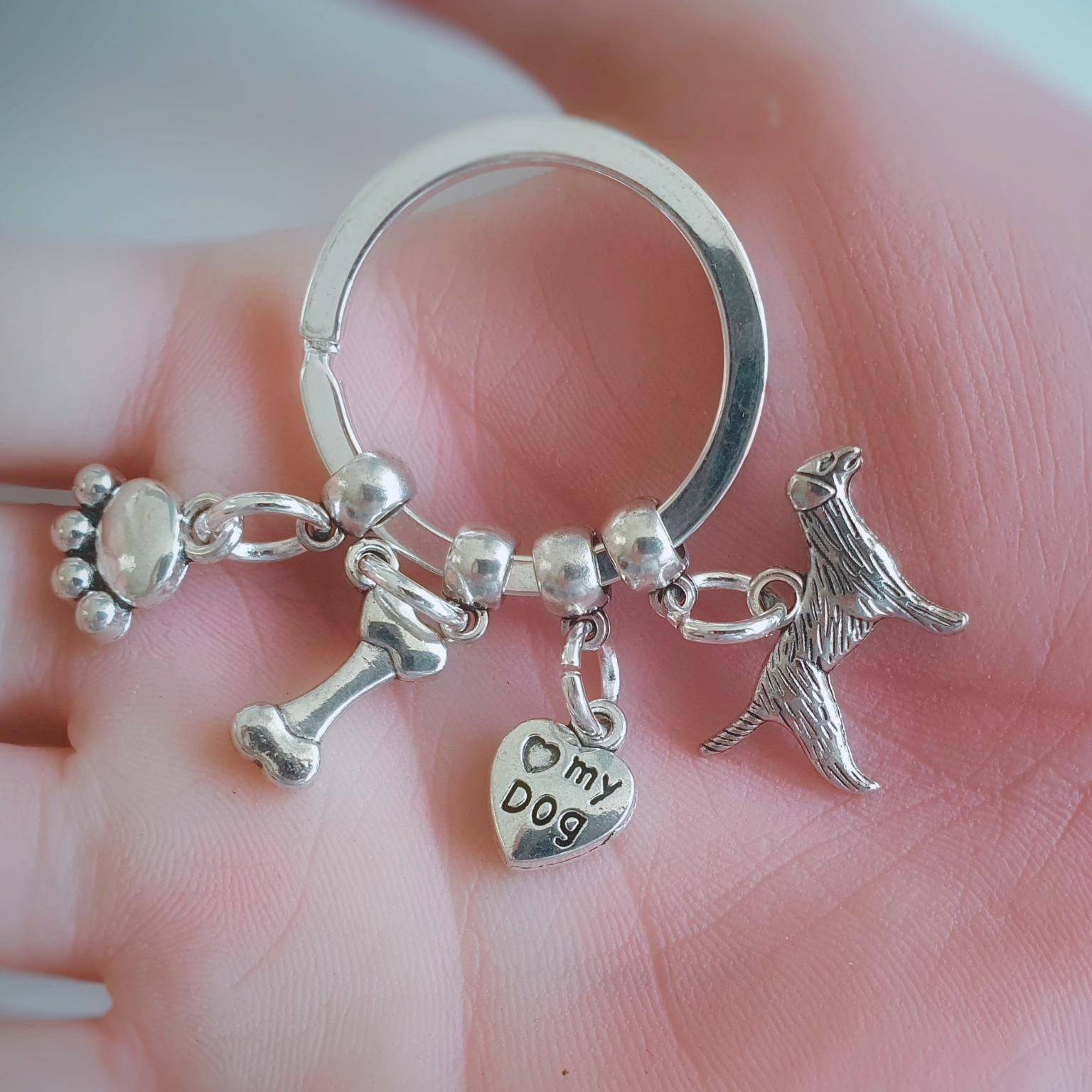 A silver-tone key ring charm set held in a person's hand. The set includes a paw print charm, a bone charm, a heart charm stamped with 'my dog', and a dog silhouette charm.