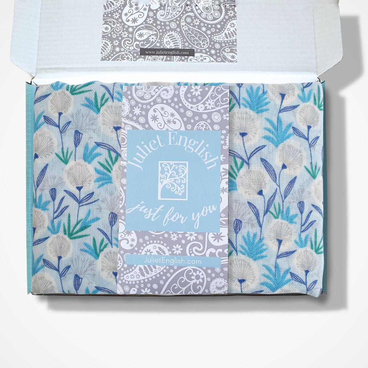 An open white gift box containing a folded patterned scarf with blue, gray, and white floral and paisley designs. A central card reads 'Juliet English Just for you' with a small logo above it.