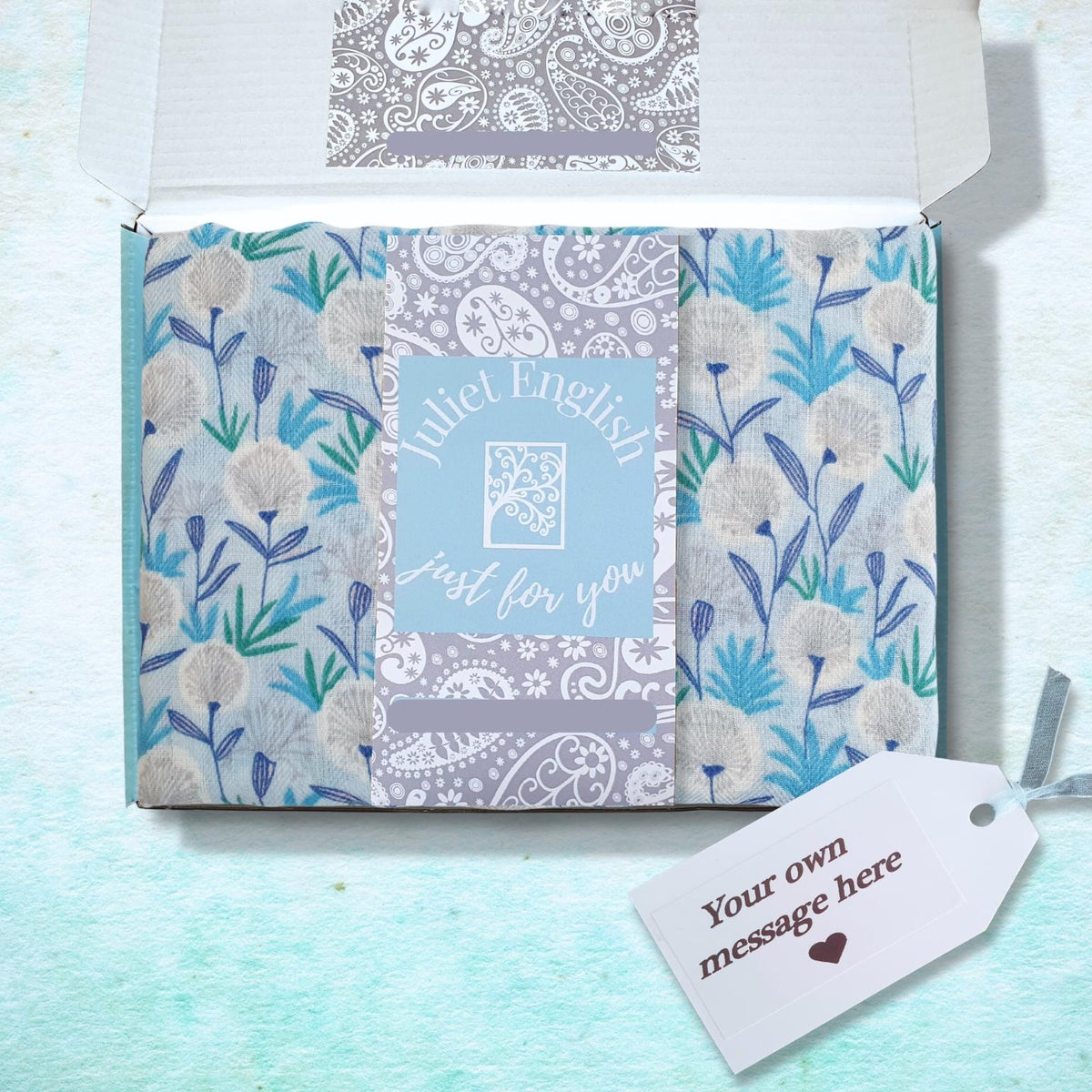 A gift box with floral scarf a in shades of blue, gray, and white, featuring a central card that reads 'Juliet English just for you'. An open white lid is visible behind it, and a small gift tag with the text 'Your own message here' lies to the right on a textured light blue surface.