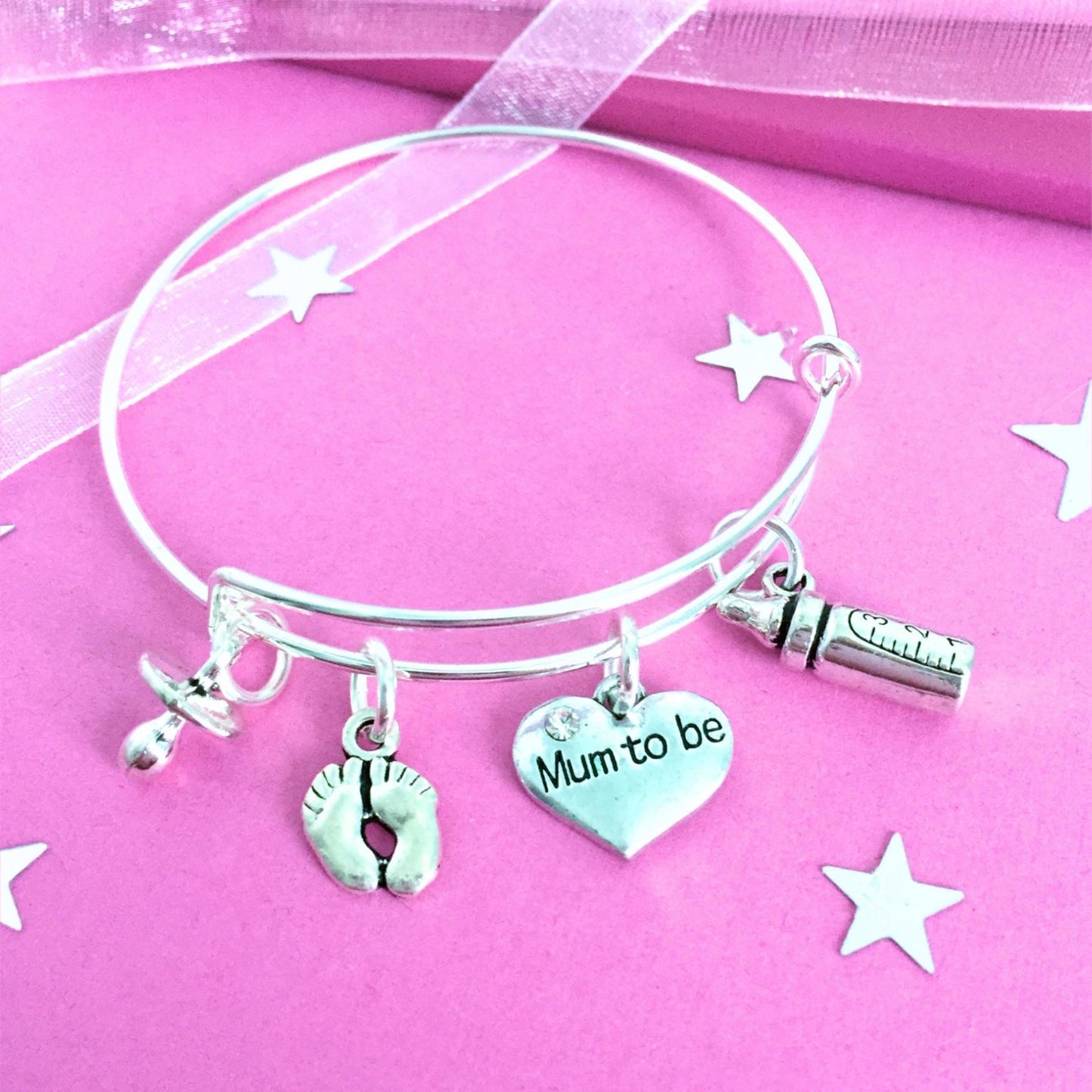 A silver wire charm bracelet with four charms: a baby footprint, a heart engraved 'Mum to be', a baby bottle, and a pacifier, displayed on a pink background with star cutouts.