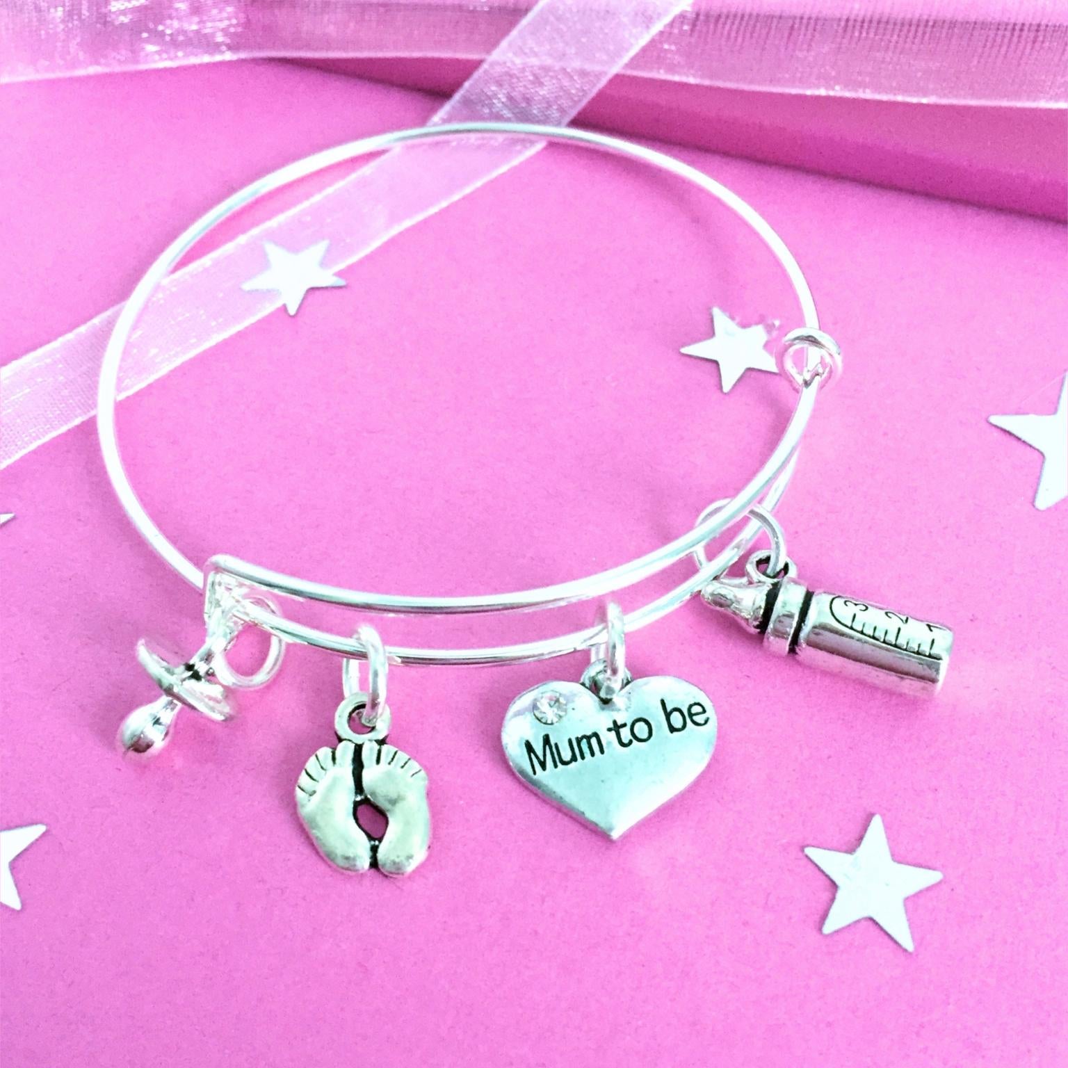 A silver wire charm bracelet with four charms: a baby footprint, a heart engraved 'Mum to be', a baby bottle, and a pacifier, displayed on a pink background with star cutouts.