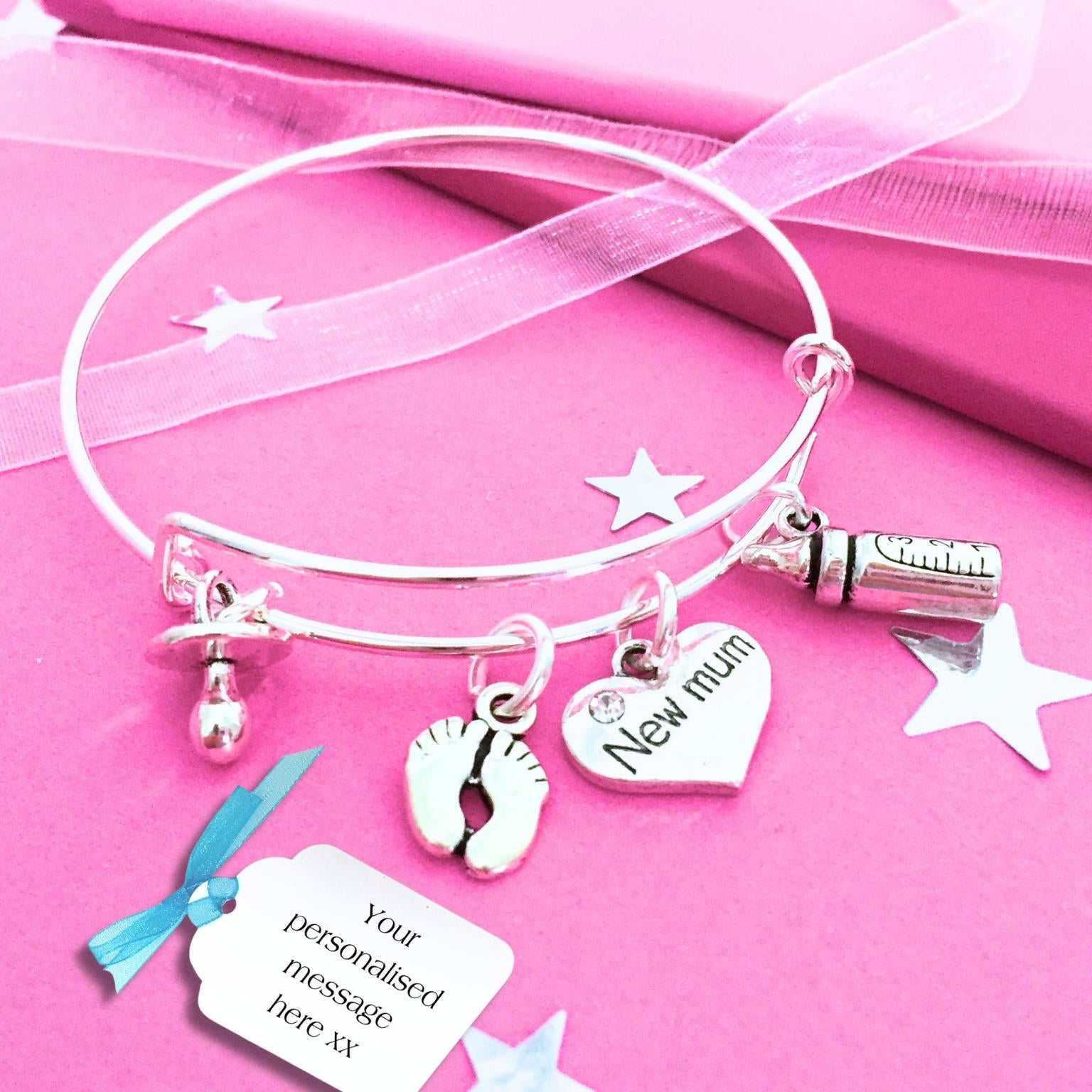 A silver-toned bangle bracelet with charms, displayed on a pink background  and stars. The bracelet includes a pacifier charm, a baby foot print charm, a heart charm engraved 'New mum', and a bottle charm.