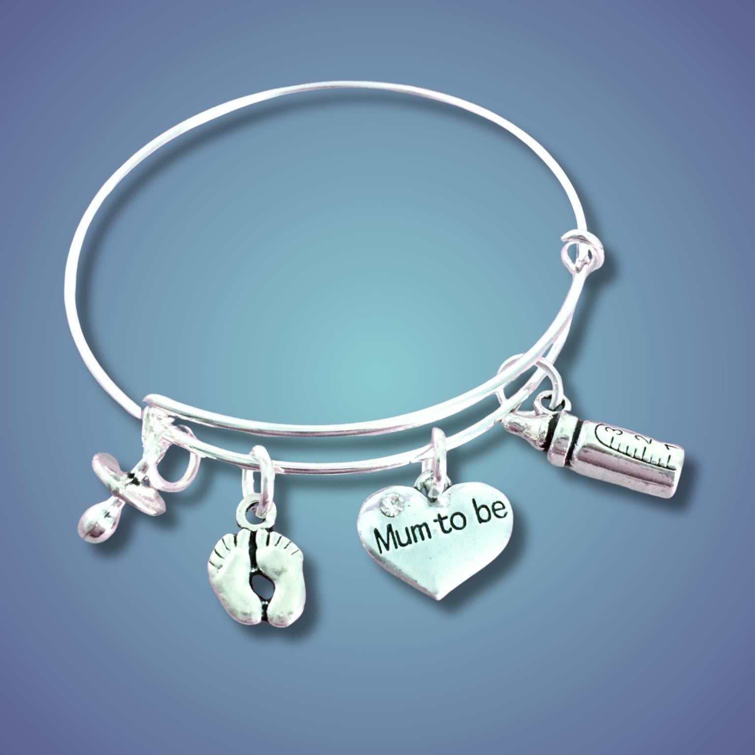 A silver bangle bracelet with four charms: a baby bottle, a pacifier, a pair of baby footprints, and a heart charm engraved with 'Mum to be', shown on a blue gradient background.